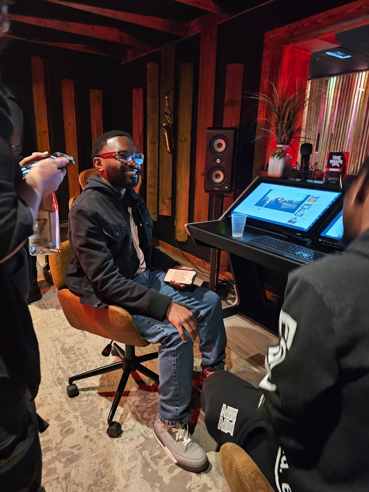 Person sitting on a chair in a recording studio, smiling, with a laptop and audio equipment nearby.