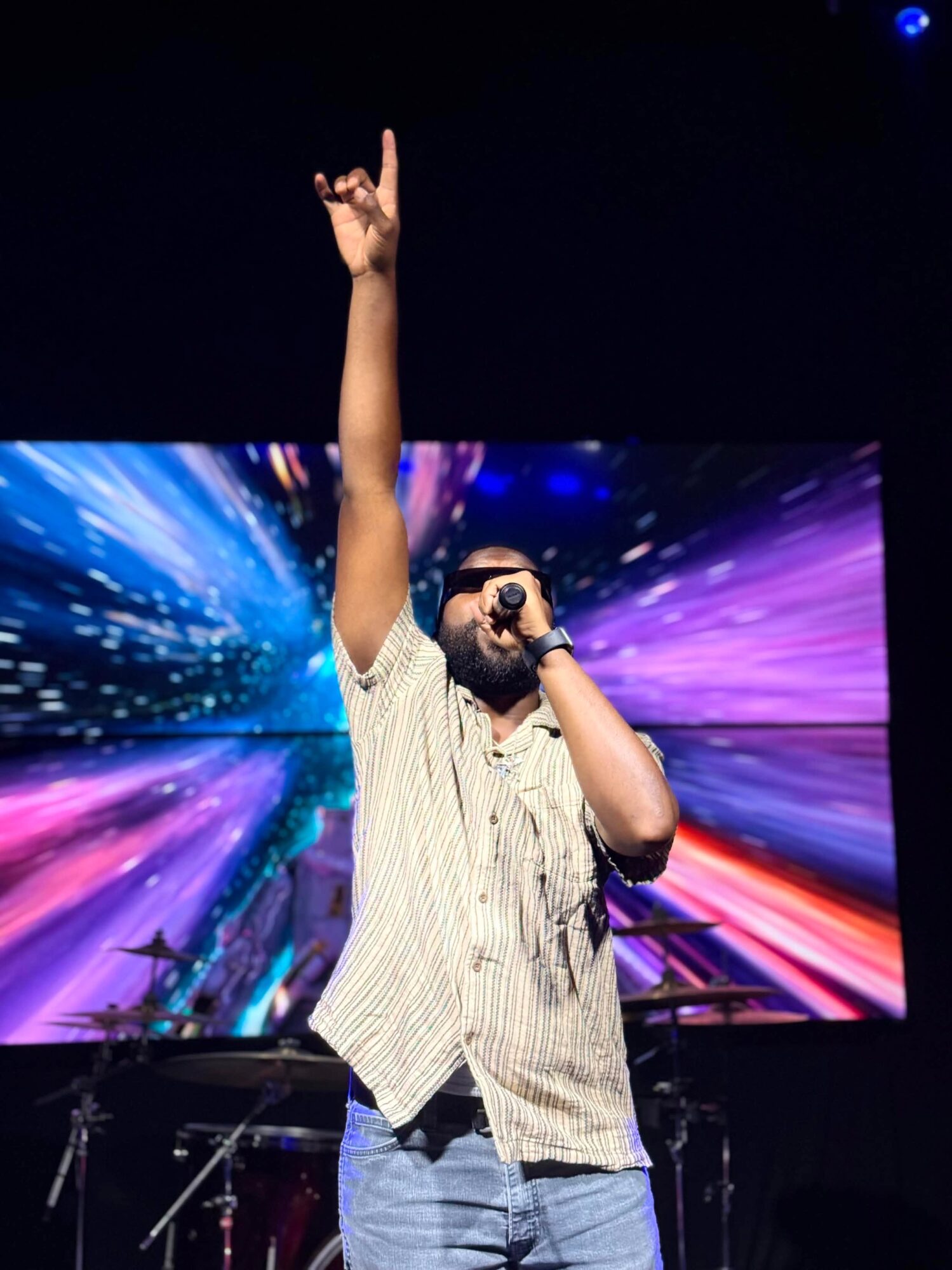 Person on stage with one arm raised, holding a microphone, in front of a colorful digital background.