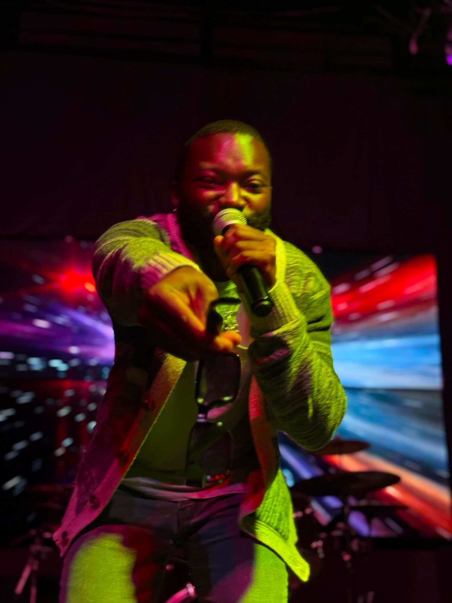 Person singing into a microphone on stage with colorful lights in the background.