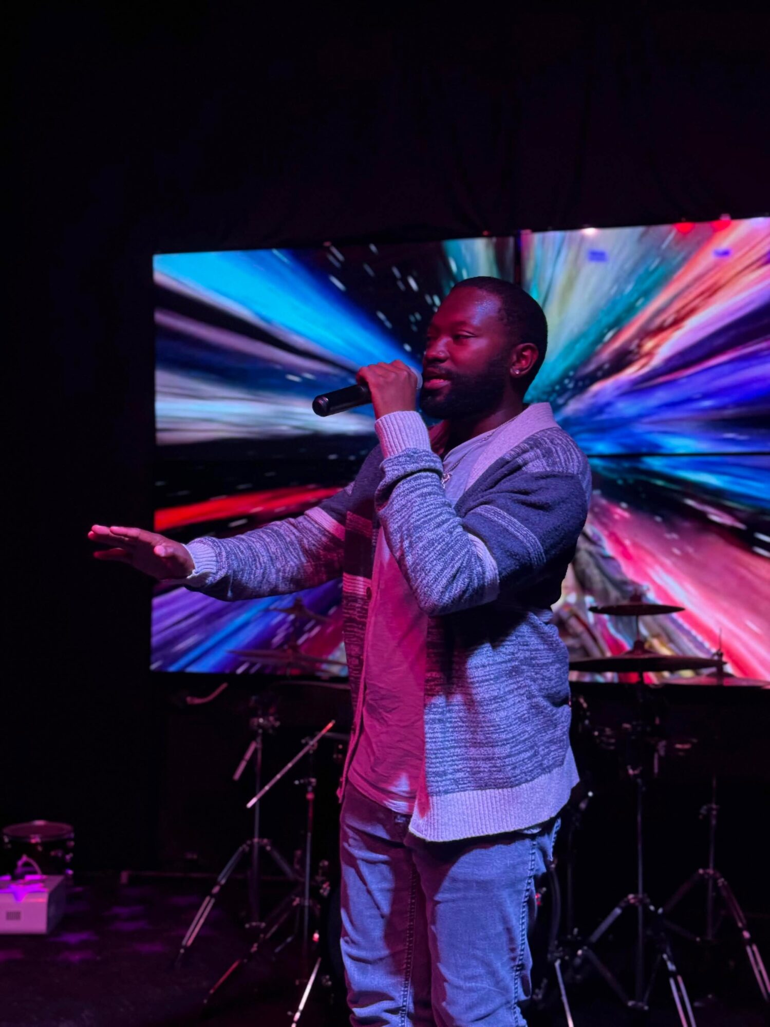 Man singing into a microphone on stage with colorful abstract background, wearing a gray jacket and jeans.