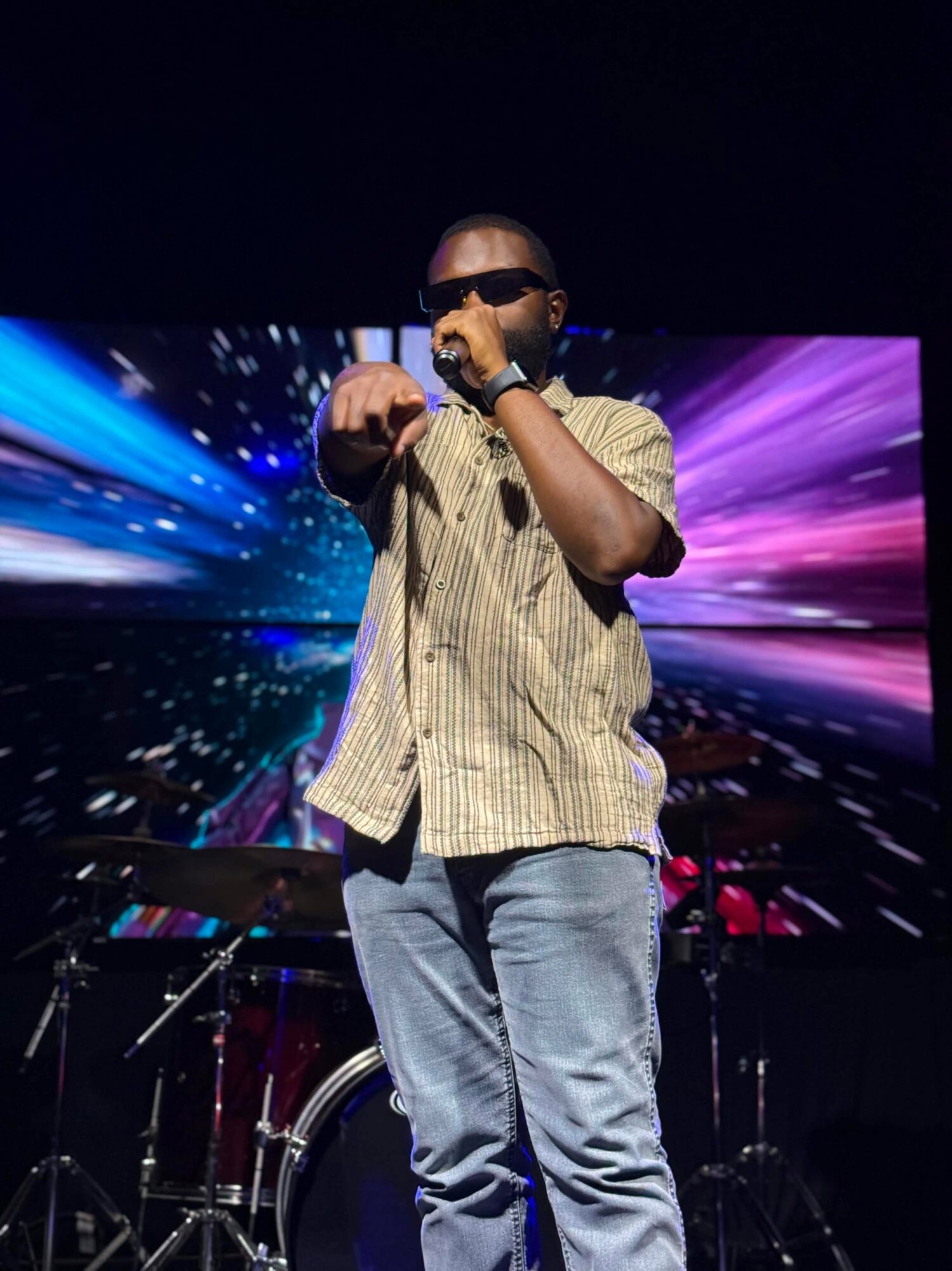 Person wearing sunglasses and a striped shirt, holding a microphone, performing on stage with colorful background display.