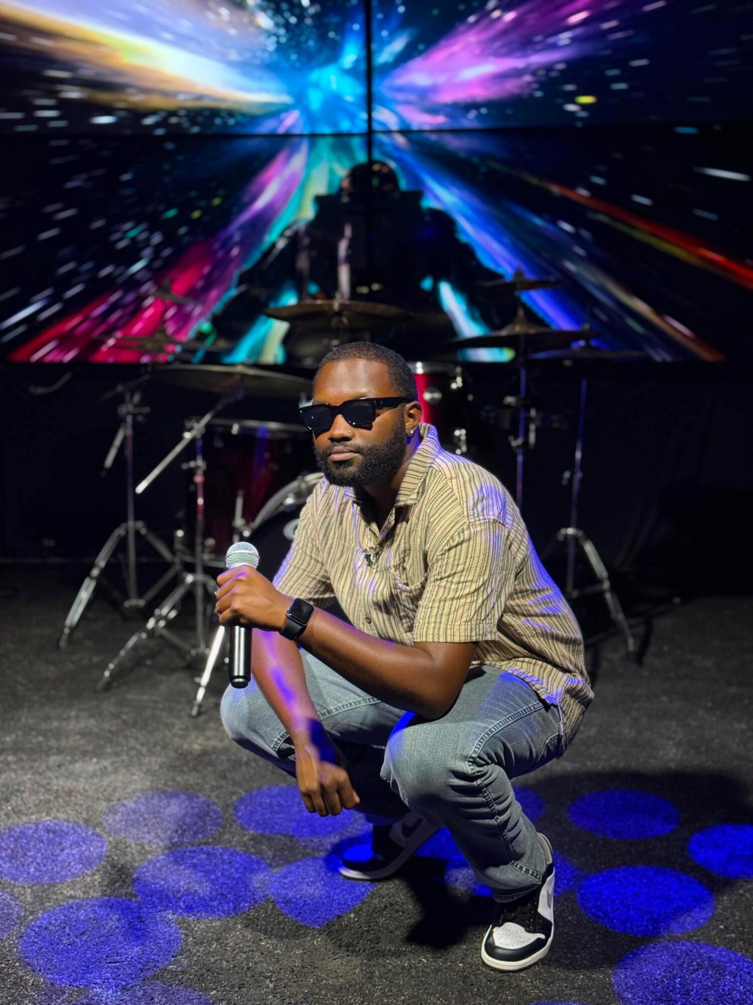 Man crouching with microphone, stage with drums and colorful digital background behind him.