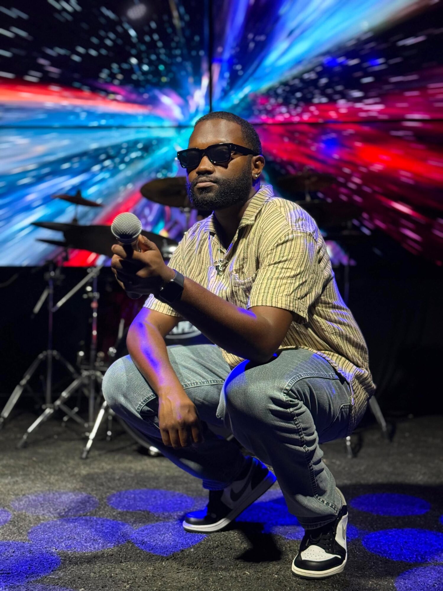 Man crouching with microphone, wearing sunglasses, in front of colorful light streaks background.