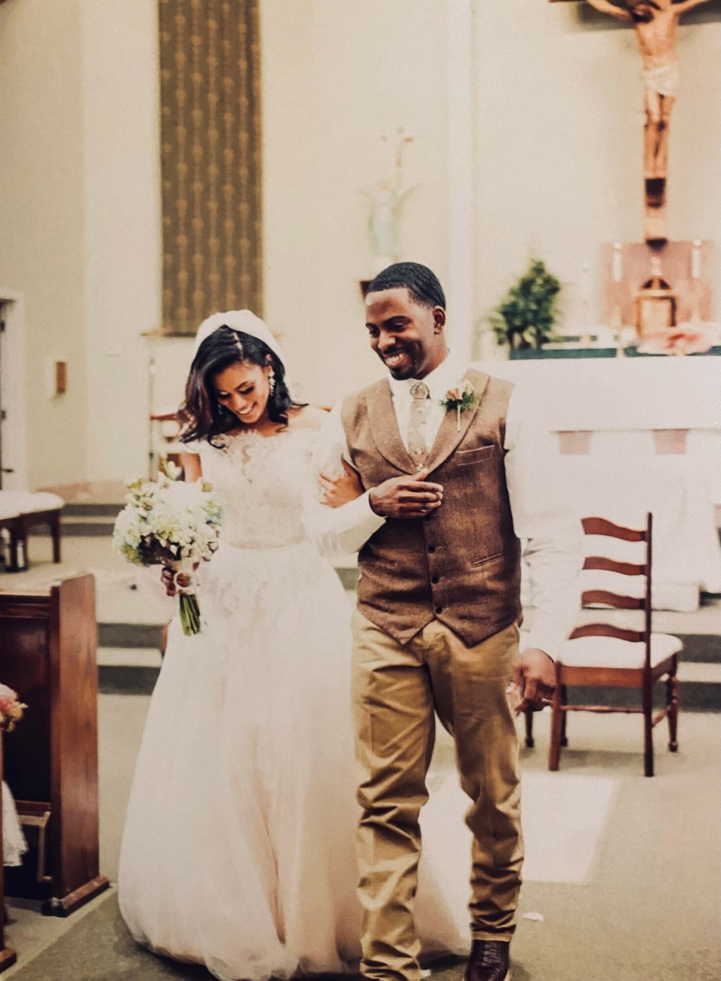Bride and groom walking down the aisle in a church, smiling, with wedding attire and floral bouquet, interior background.