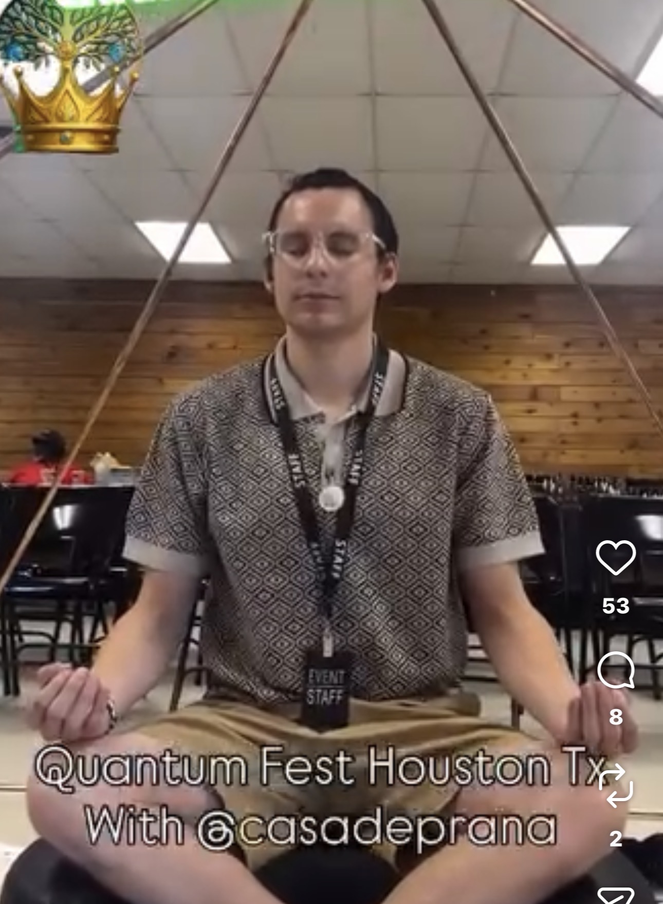Person sitting cross-legged with eyes closed, meditating indoors with wooden wall and ceiling, crown decoration above, and a sign reading 'Quantum Fest Houston Tx' below.