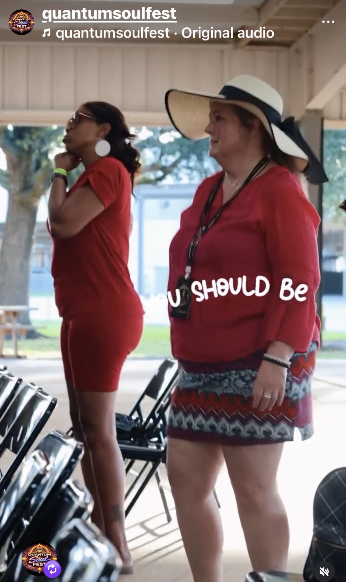 Two women standing outdoors, one in a red dress and the other in a red top with a patterned skirt and large hat, talking.