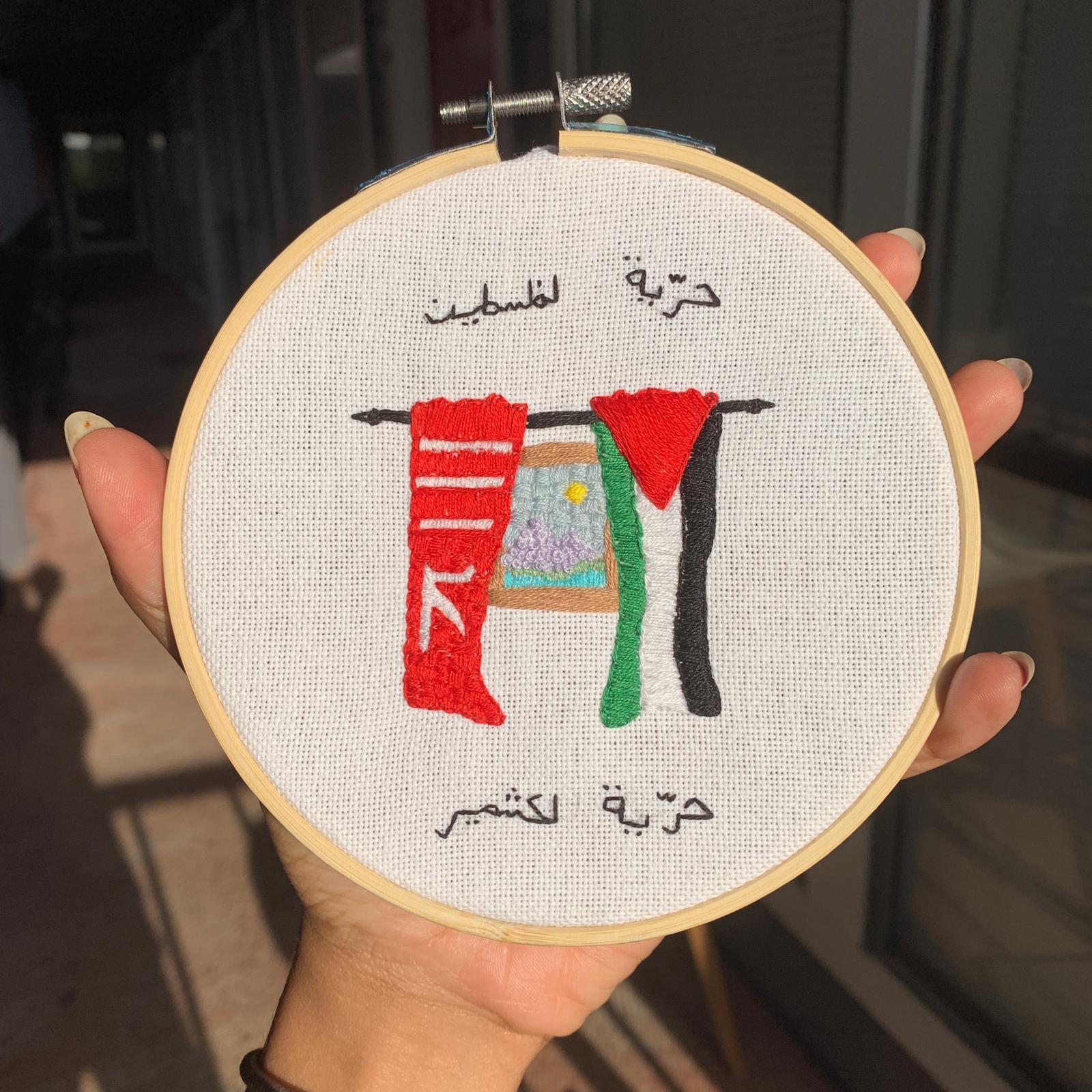 Embroidery hoop with clothes hanging, Arabic text above and below, person holding it outdoors, sunlight, hand visible.