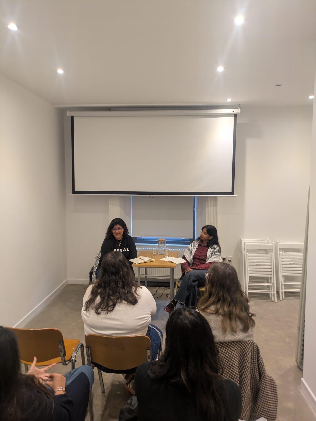 Group of five women sitting in a room with a large screen and stacked chairs, engaged in a discussion.