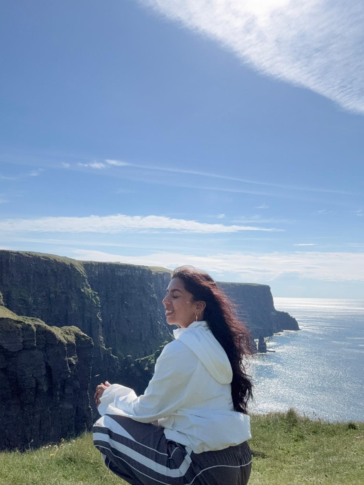 Woman with long dark hair sitting on grass near cliffs and ocean under blue sky.
