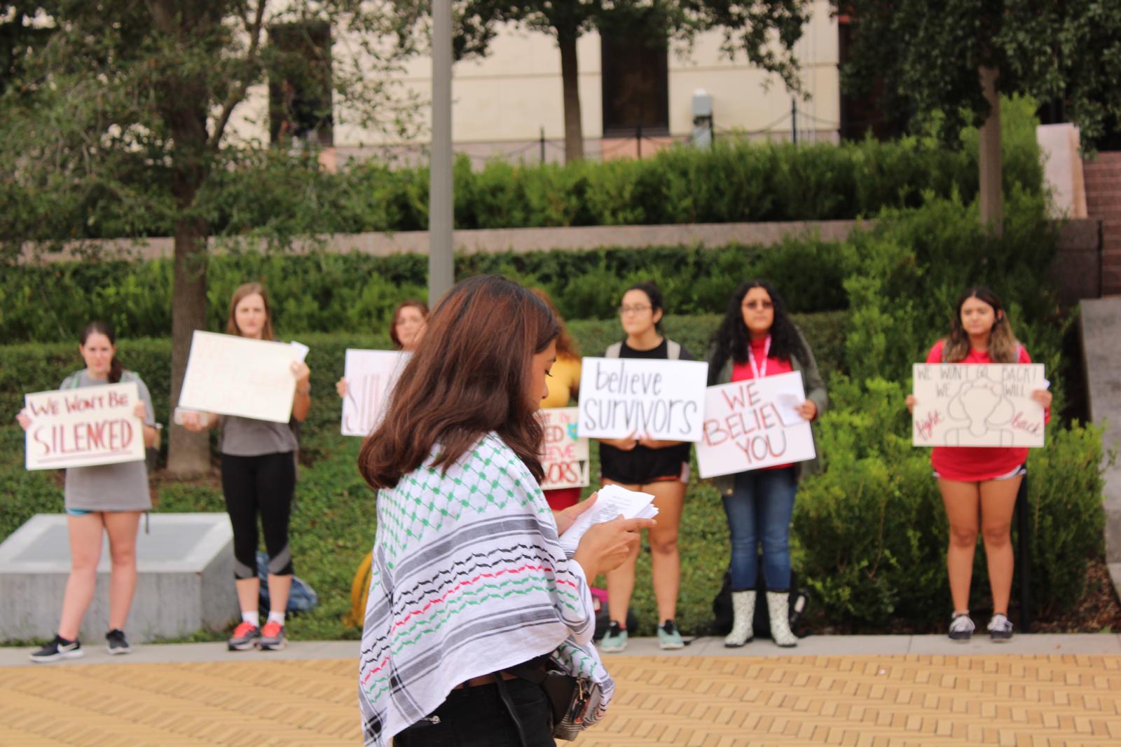 Group of women protesting outdoors, holding signs, with trees and bushes in the background.