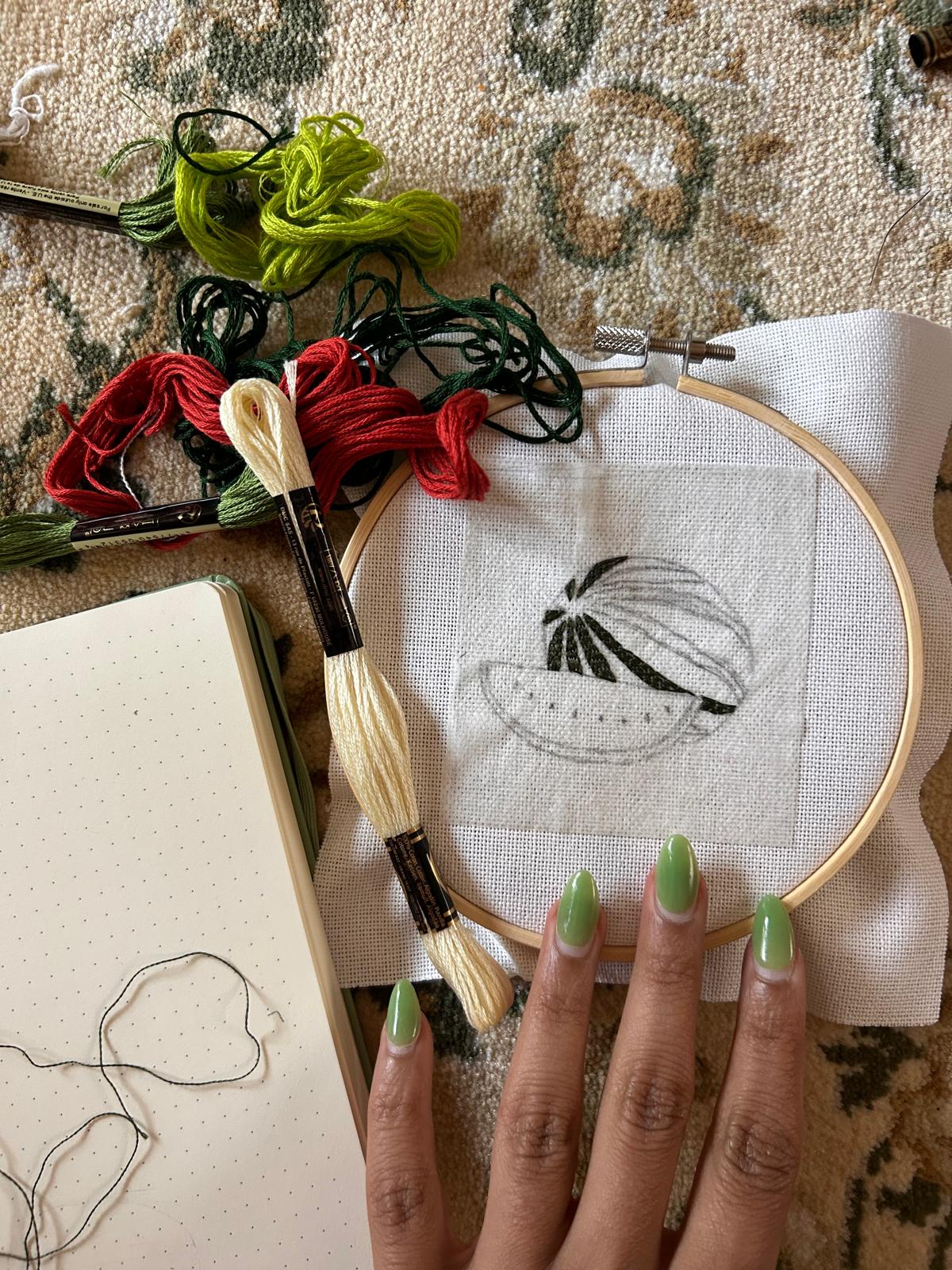 Embroidery hoop with a design, embroidery floss, and a person's hand with green nails on a textured surface.