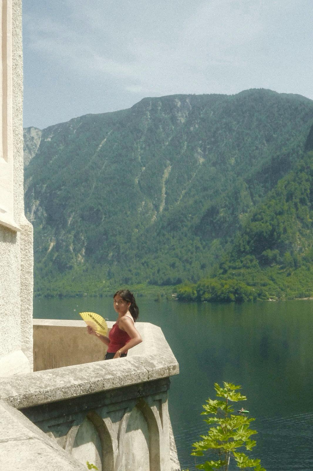 Girl reading a book on a balcony overlooking a lake and mountains, with a small tree in the foreground.