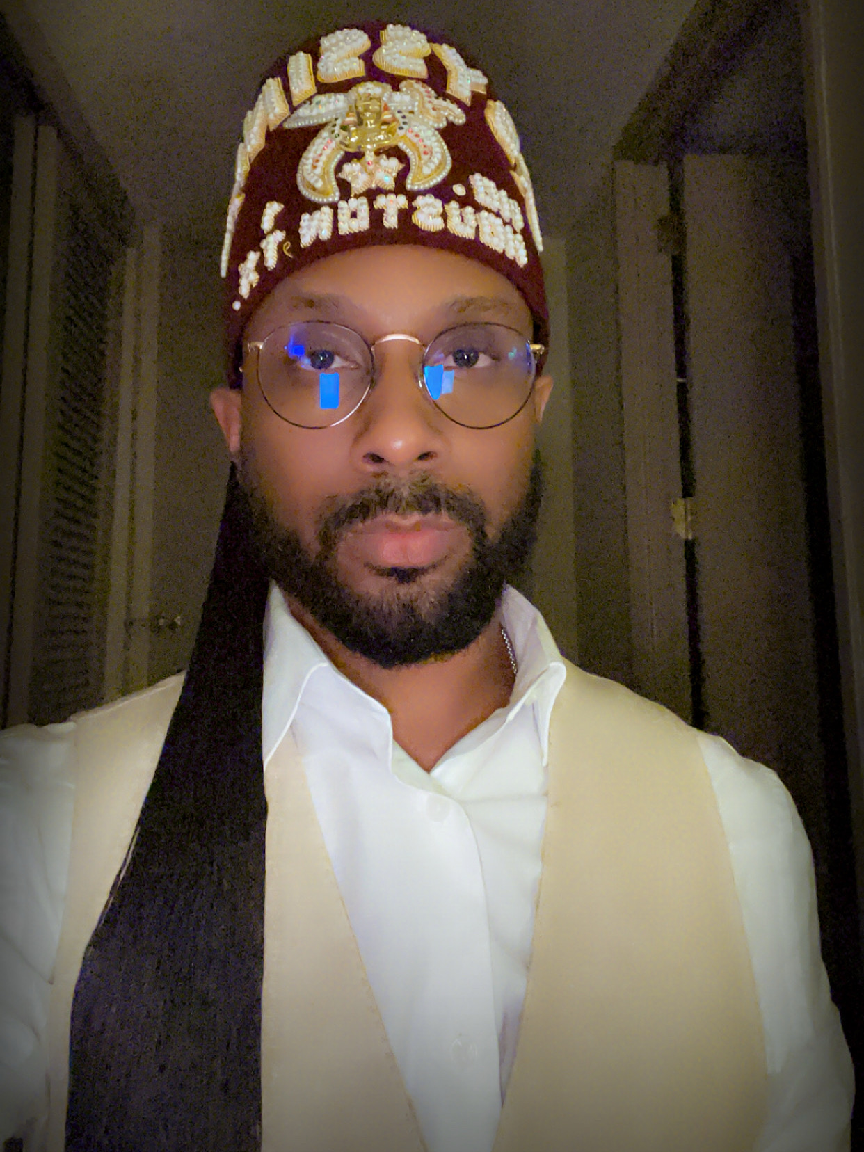 Person wearing a red and gold ceremonial hat, glasses, white shirt, and cream vest, indoors at night.