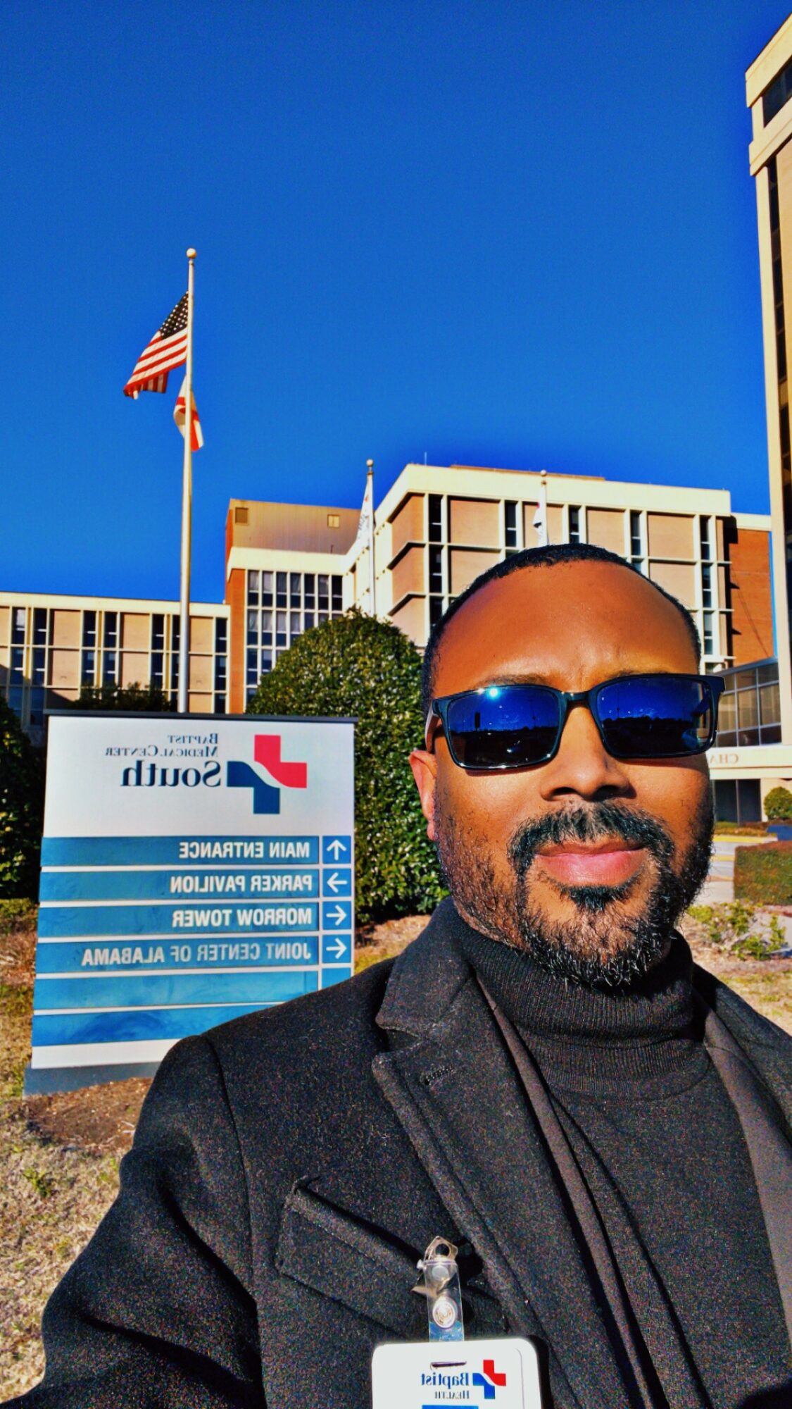 Man with sunglasses and beard taking a selfie outside a hospital building, blue sky background.
