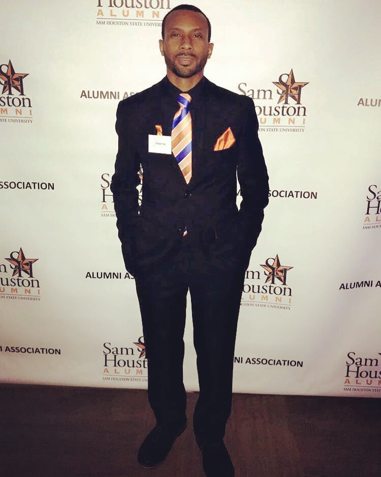 Man in a suit standing in front of a backdrop with logos, hands in pockets, facing forward.