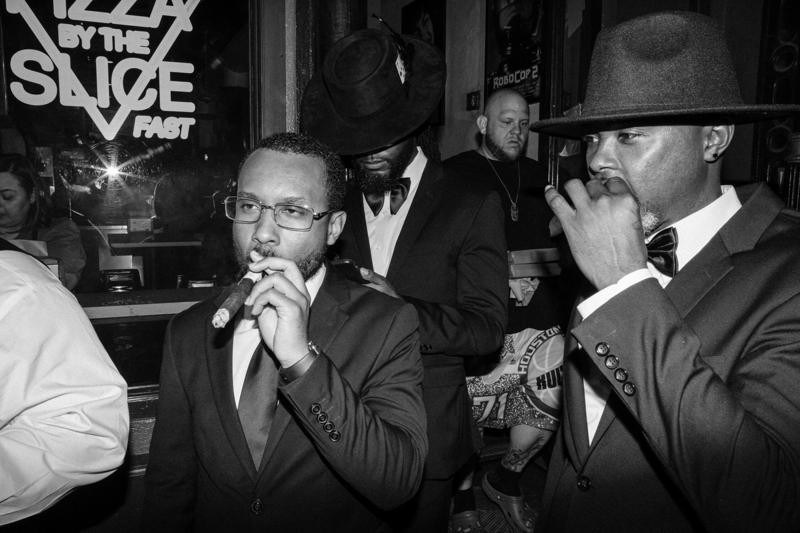 Group of men in suits and hats, some holding microphones, in a dimly lit indoor setting, possibly a bar or club.
