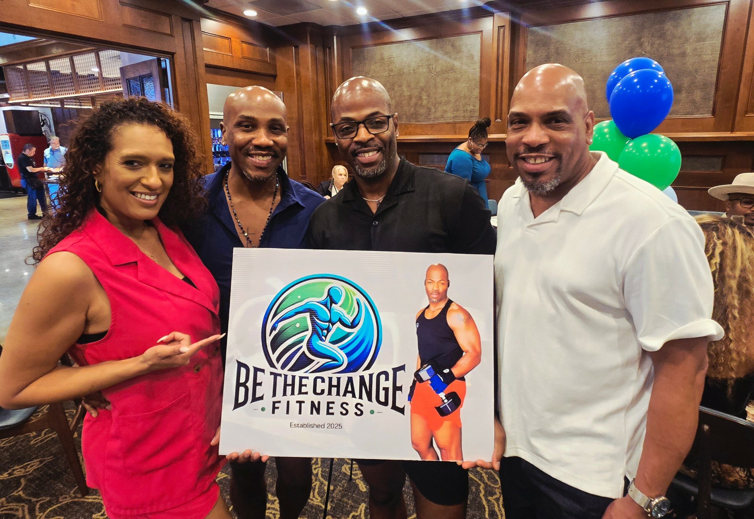 Four people holding a fitness sign, smiling indoors with balloons in the background.