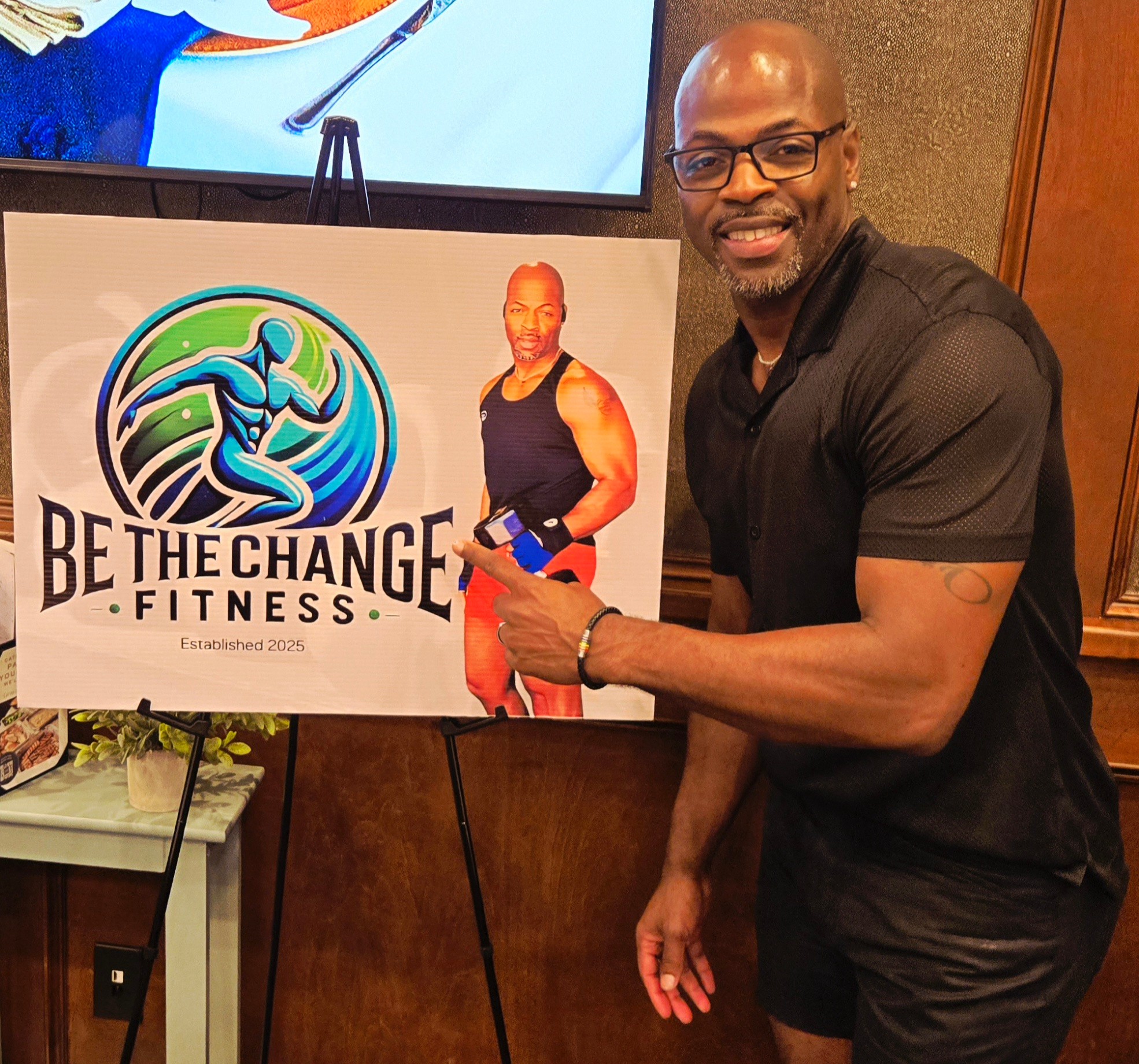 Man with glasses and a beard points to a fitness poster with a woman in workout clothes, smiling at the camera.
