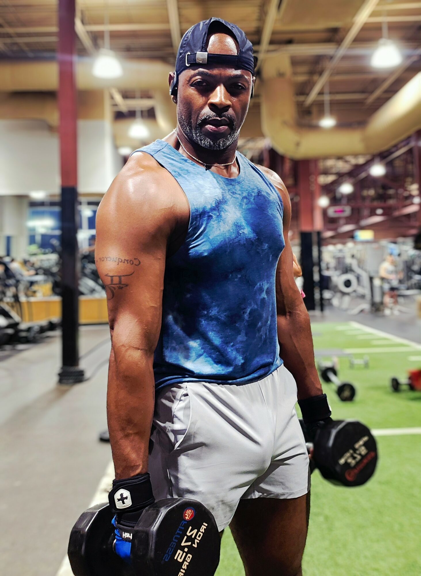 Man in gym holding dumbbells, wearing a blue sleeveless shirt, gray shorts, and a cap, with gym equipment in background.