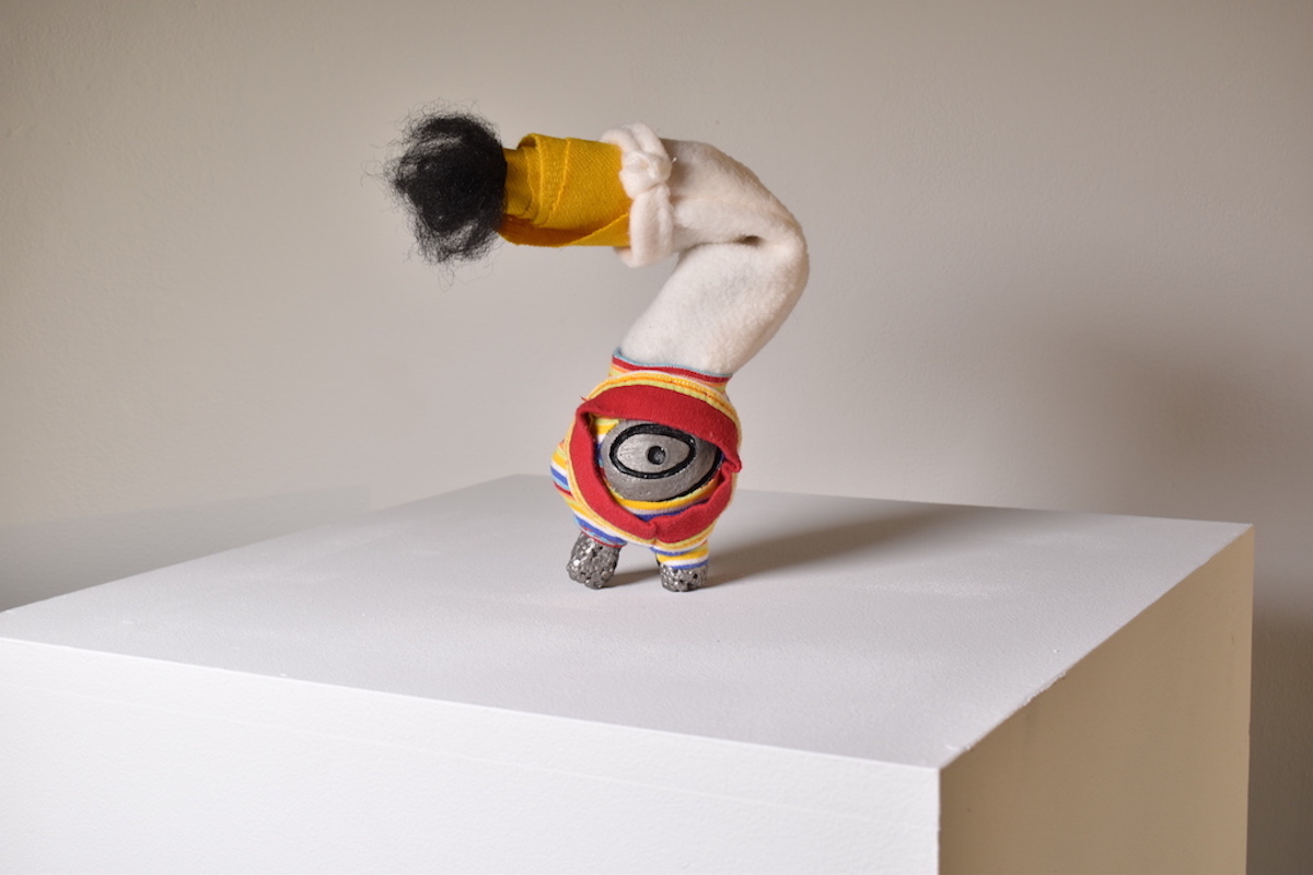 Person performing a handstand with legs bent, wearing colorful clothing and a hat, on a white platform.