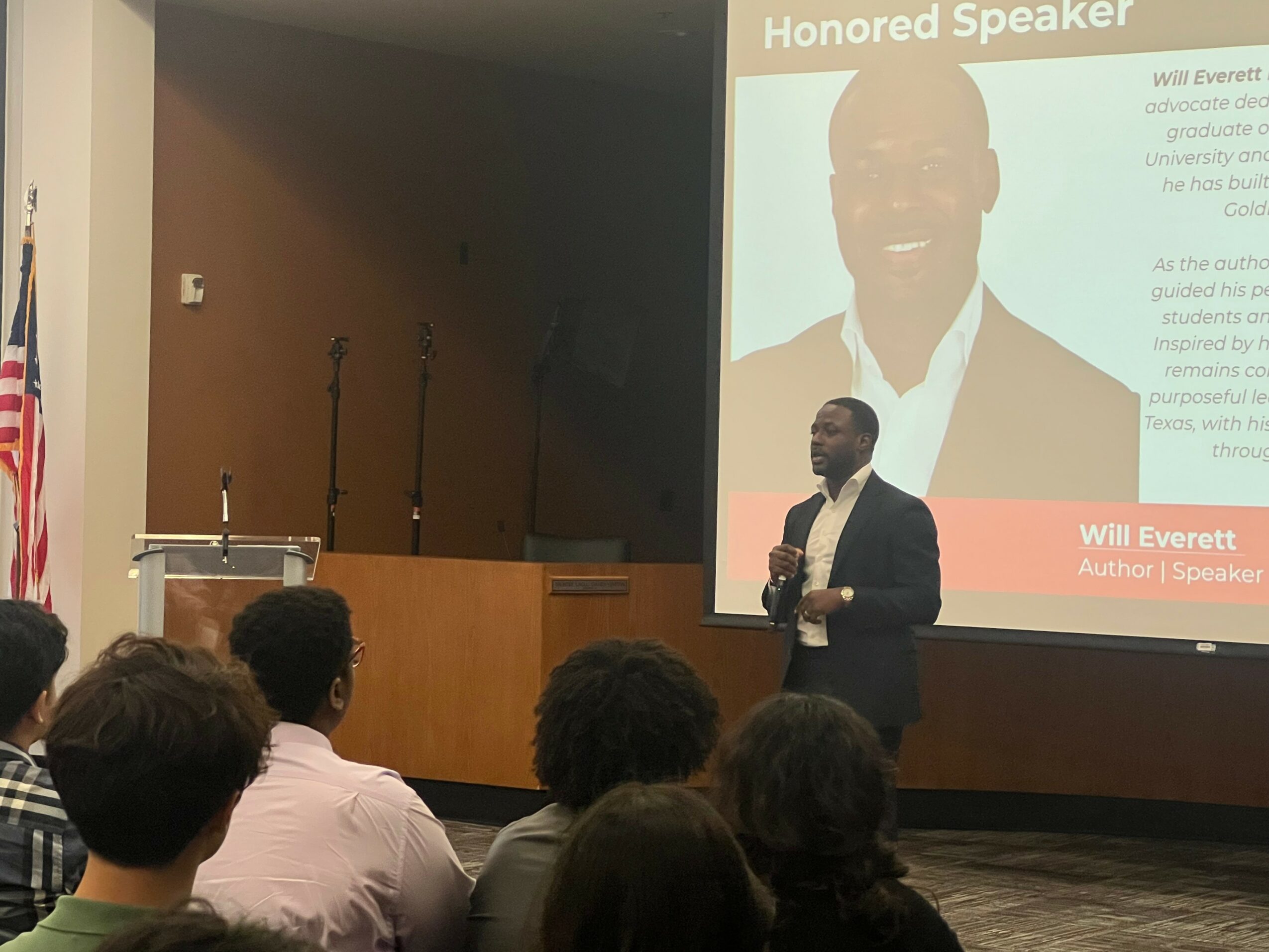 Speaker in a suit holding a microphone presents to an audience in a conference room with a large screen displaying a slide.