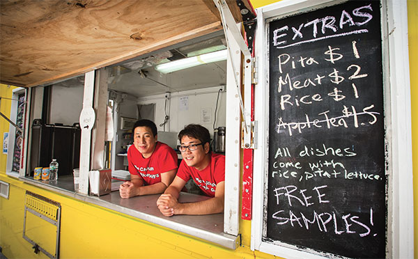 Two children in red shirts leaning on a food truck counter, smiling, with a menu board listing extras and free samples.