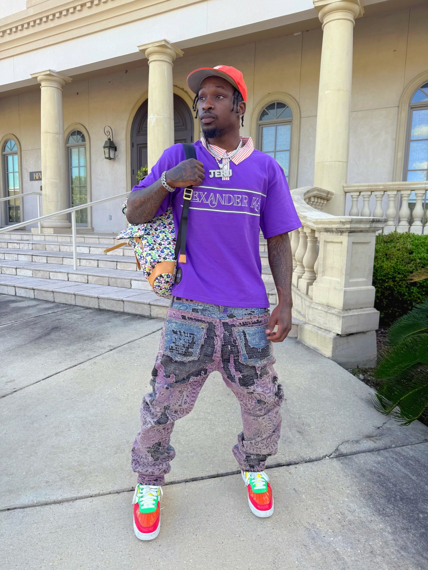 Young man standing outside a building with columns, wearing a purple shirt, patterned pants, colorful sneakers, and a red cap, holding a bag.