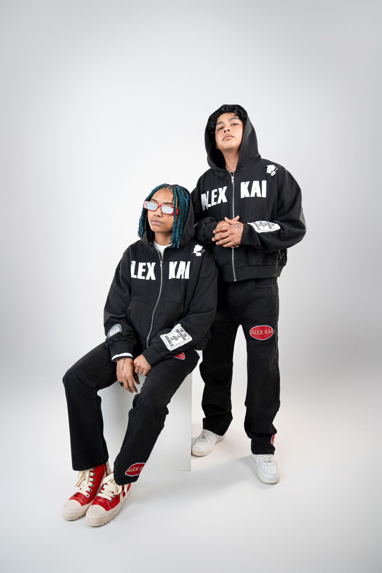 Two women in black hoodies and pants, one sitting and one standing, against a plain white background.