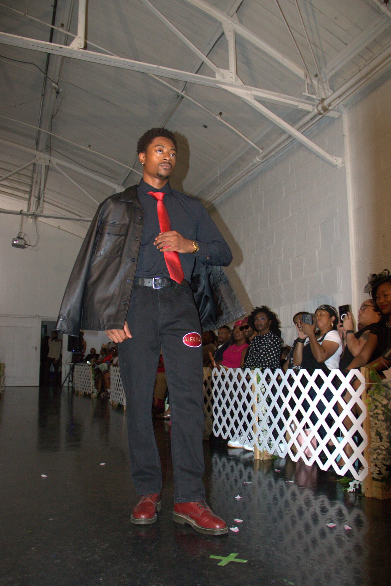 Tall man with curly hair wearing a black jacket, red tie, and black pants standing in an industrial space with audience behind a white fence.