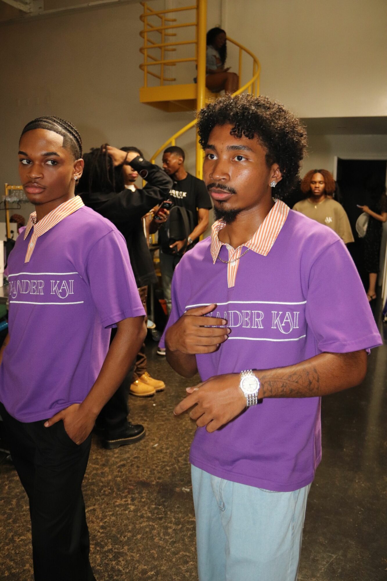 Two women wearing purple shirts with white text, standing indoors with other people in the background.
