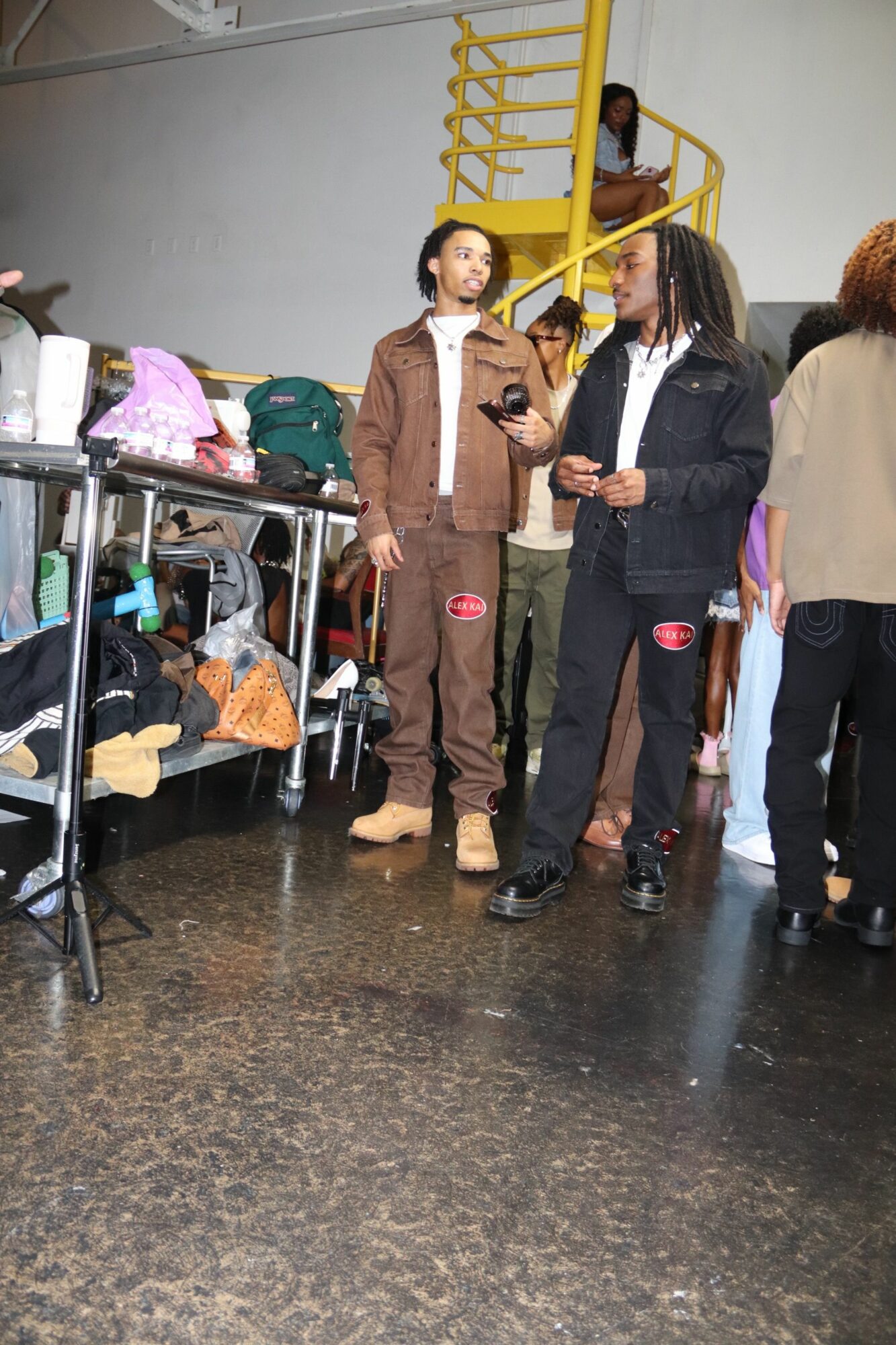 Two women and a man standing in a room with a yellow ladder and various items, some people sitting on a bench.