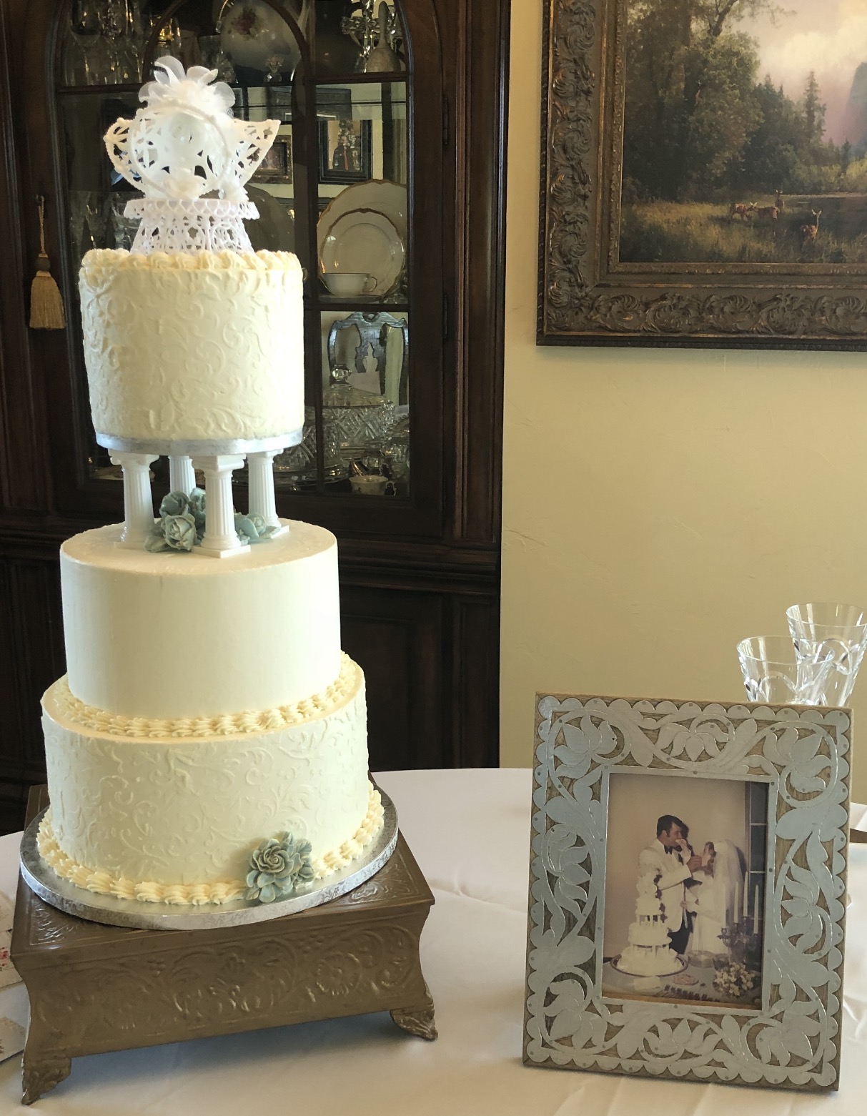Three-tiered white wedding cake with decorative flowers, topped with a floral arrangement, on a table with a framed photo nearby.