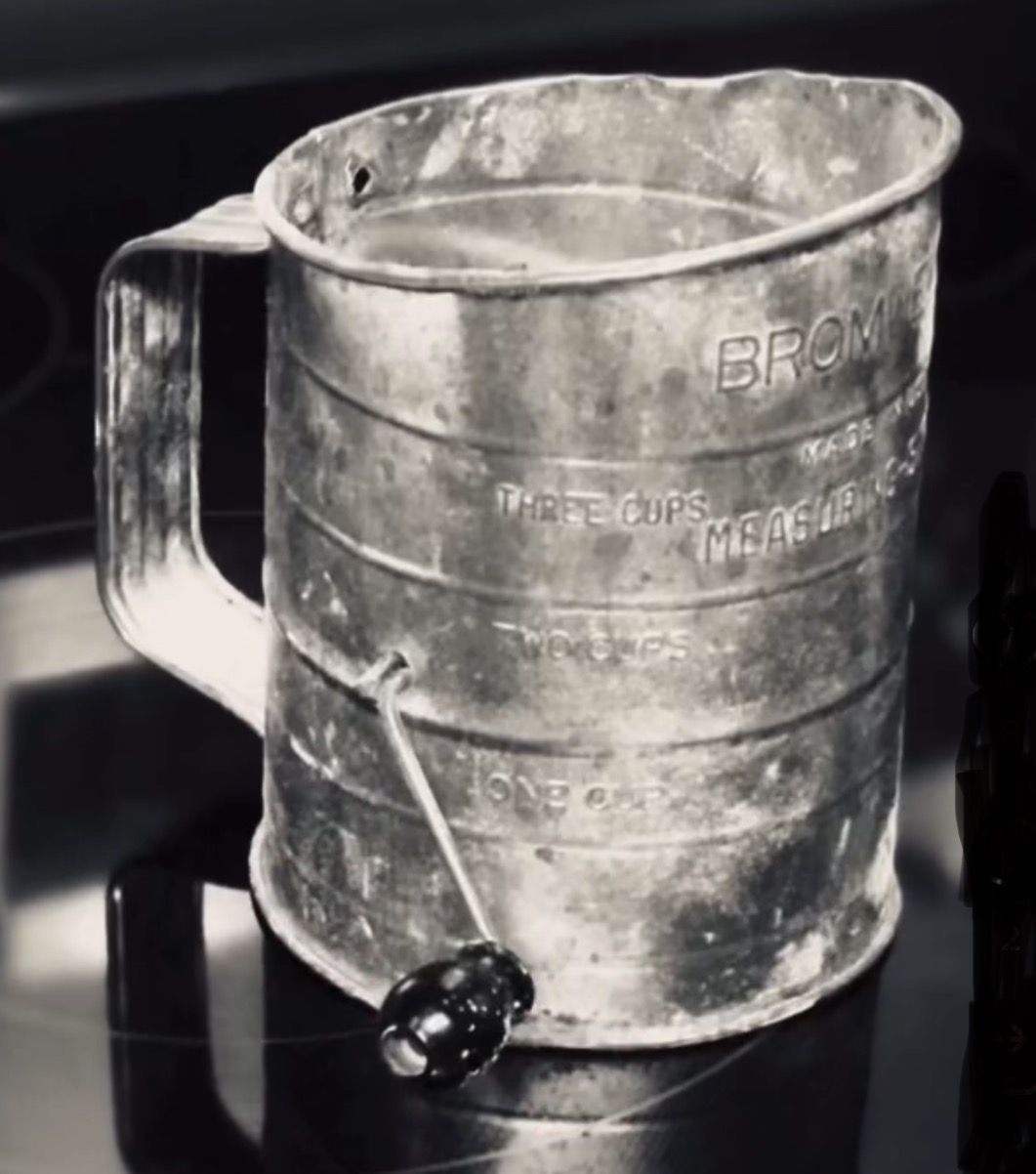 A weathered metal measuring cup with a handle, a small black object attached with a string, on a dark surface.