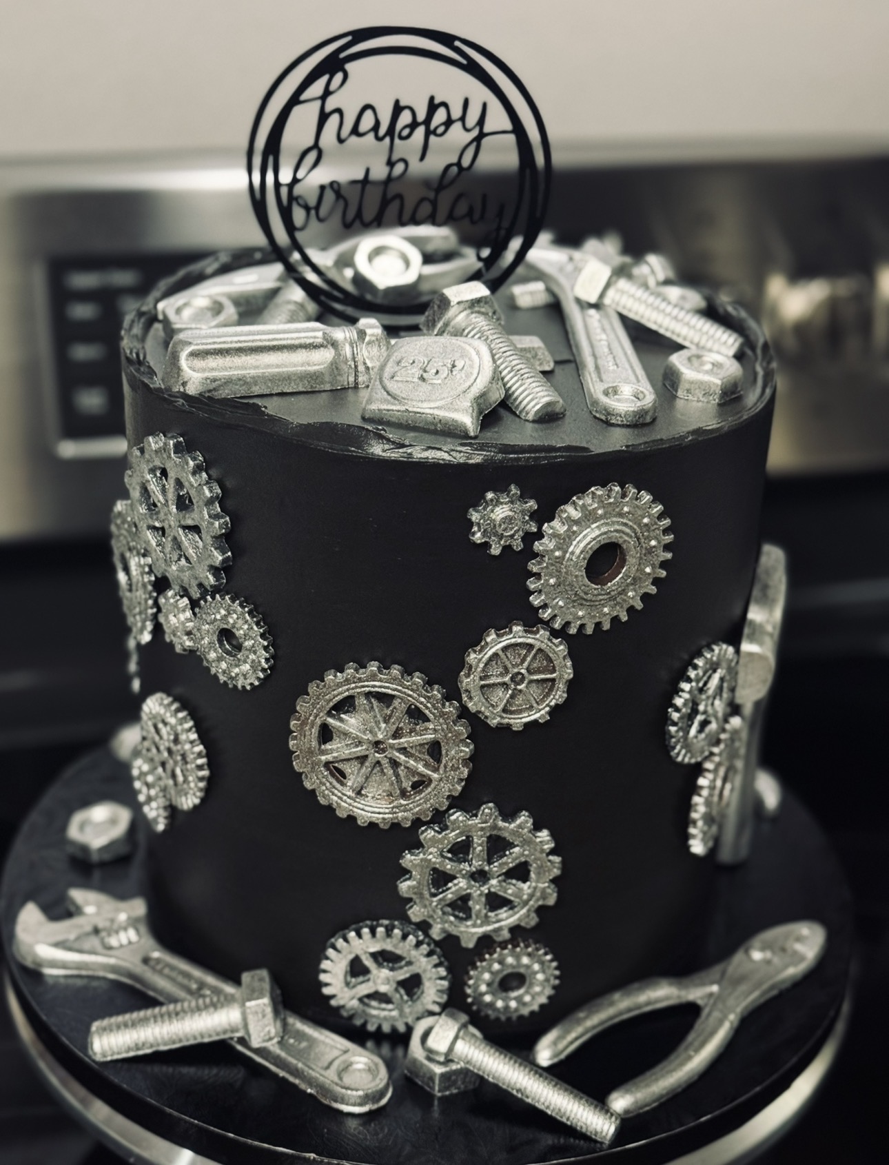 Black birthday cake decorated with silver gears, tools, and a 'happy birthday' topper, on a black base.