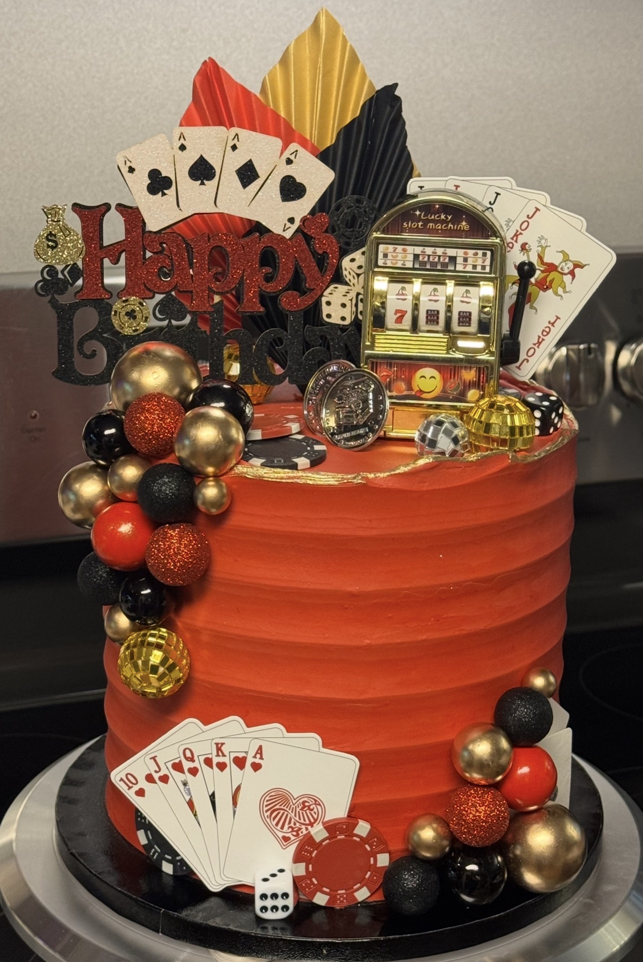 Celebration cake decorated with playing cards, poker chips, and a slot machine, topped with colorful leaves and a 'Happy' sign.