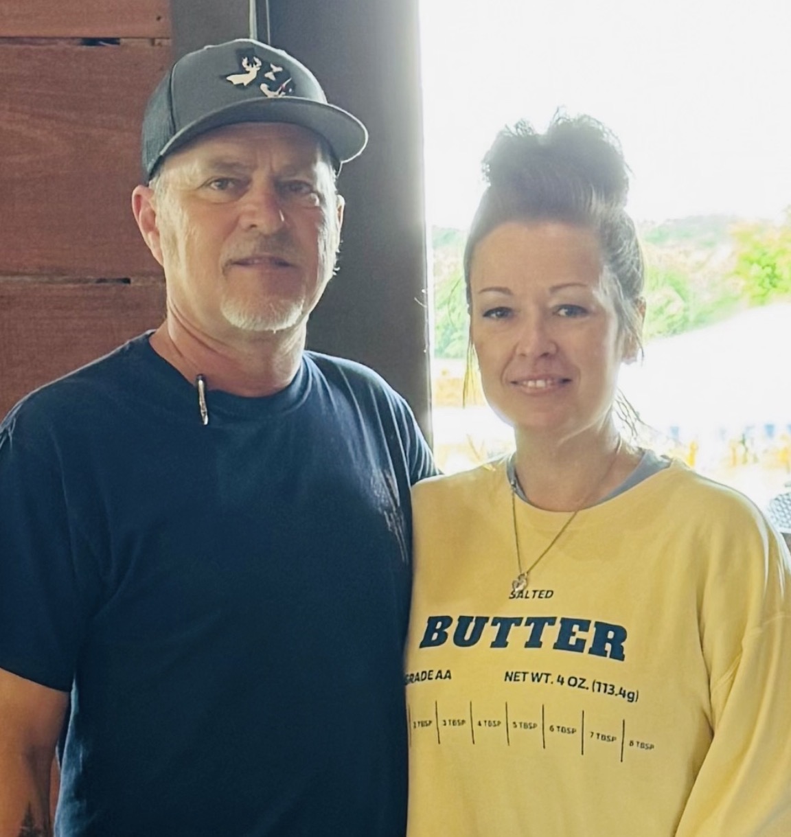 Two people standing indoors near a window, smiling, with a wooden wall behind them and bright outdoor scenery visible through the window.