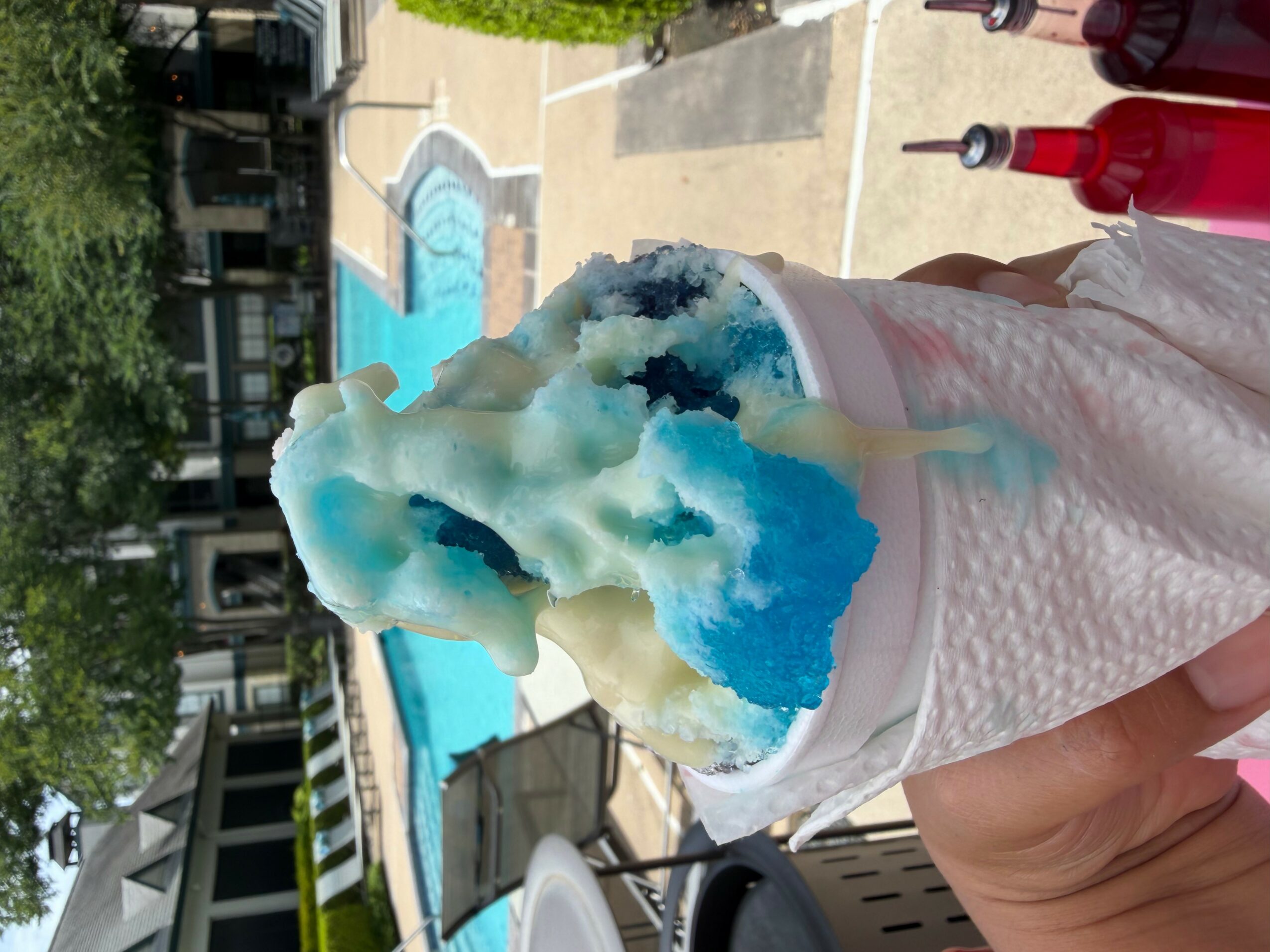 Person holding a cup of blue and white cotton candy near a swimming pool, with trees and buildings in background.