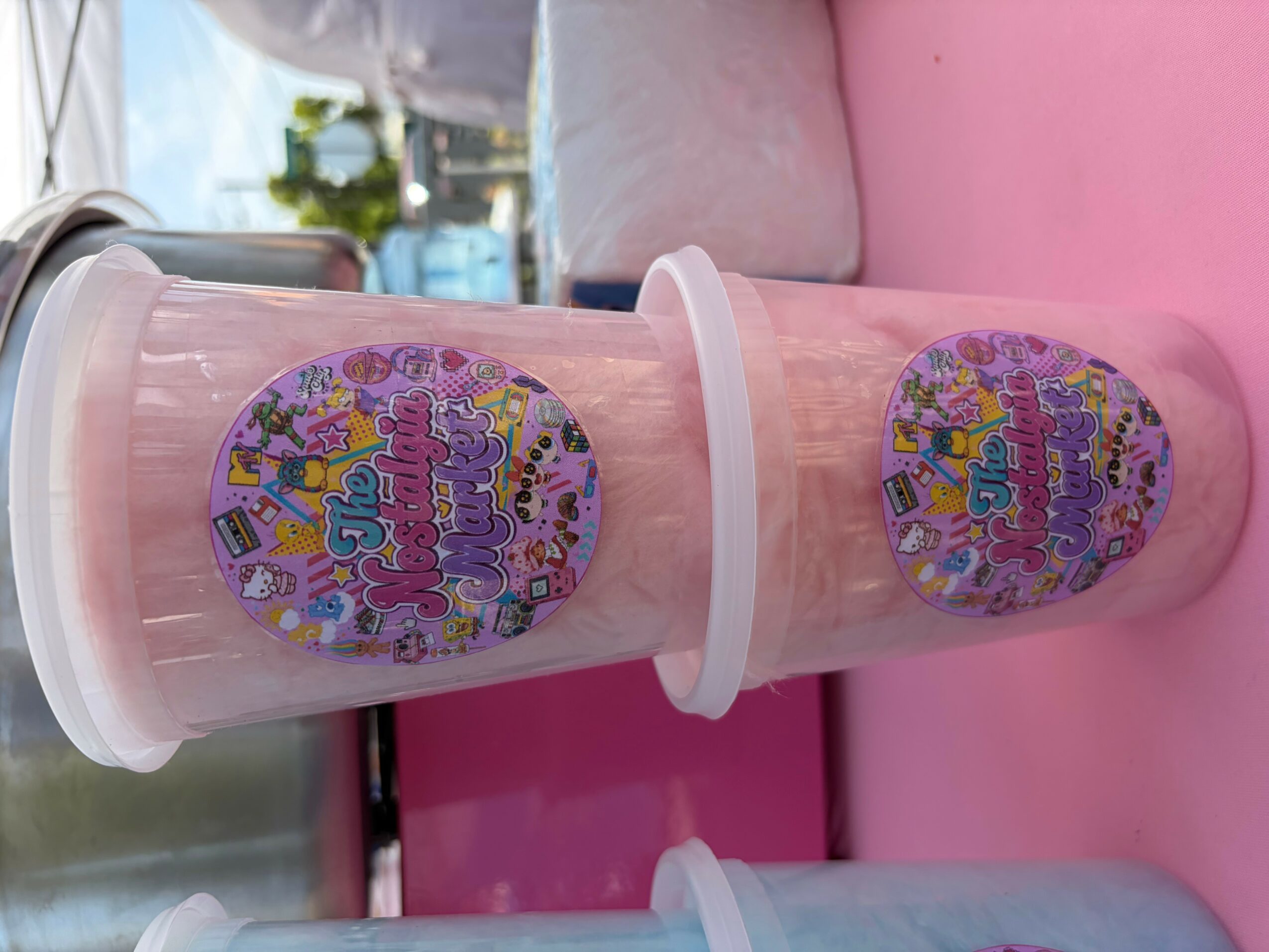 Pink milkshake in a clear cup with a lid, colorful sticker label, and pink straw, on a pink surface.