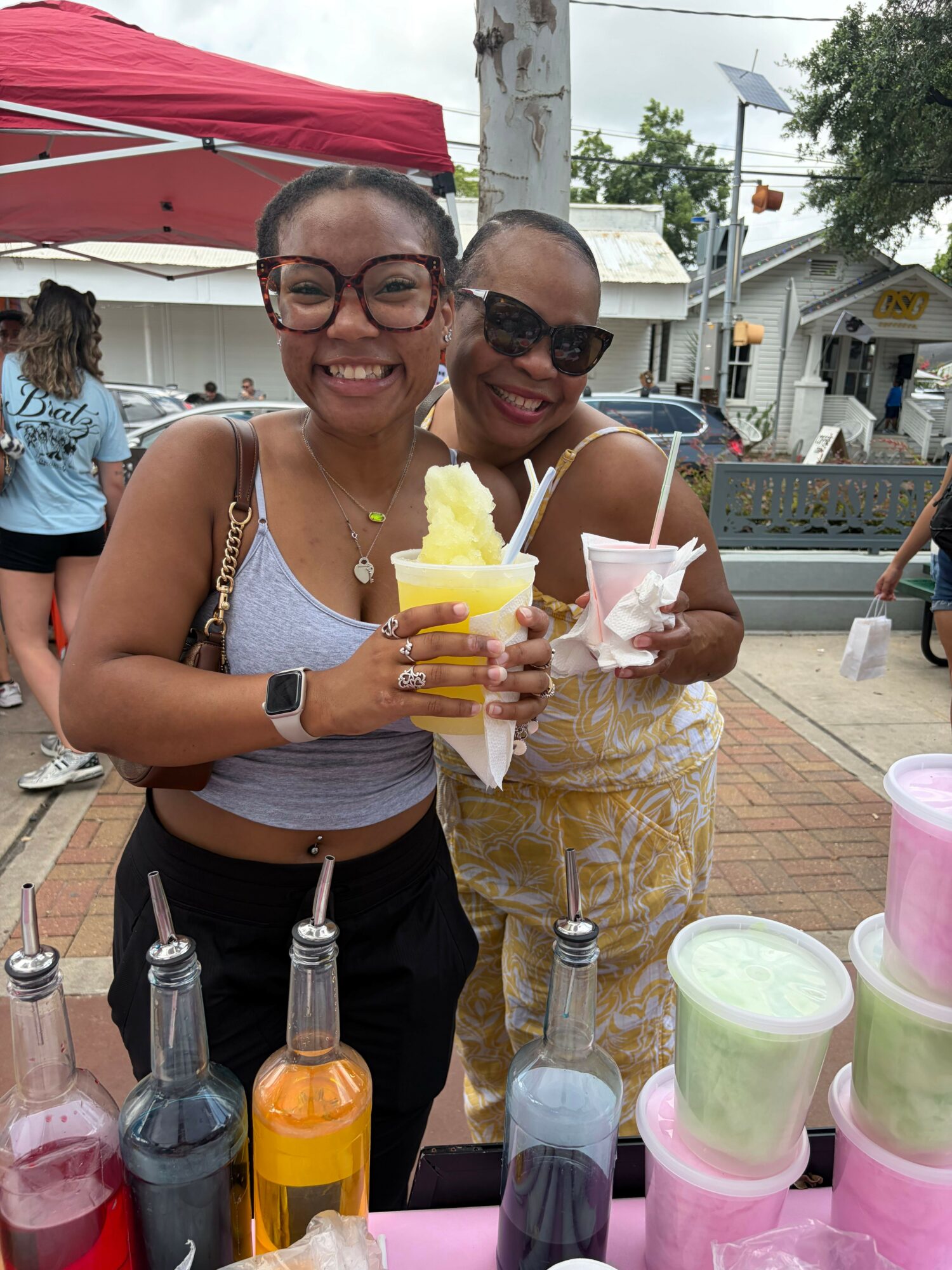 Two women at an outdoor event, one holding a yellow drink, surrounded by colorful cups and bottles, with a red tent in the background.
