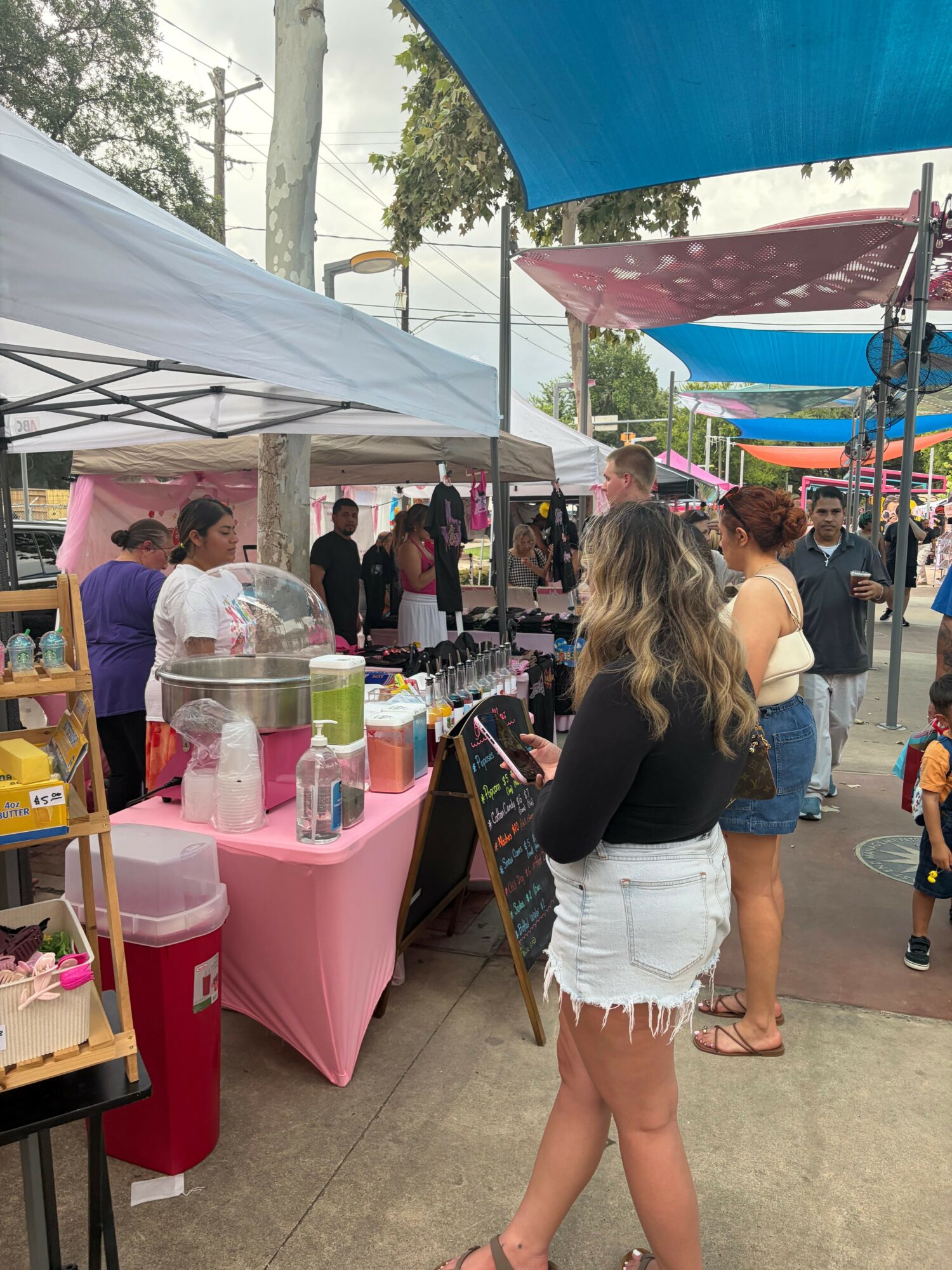 People browsing at an outdoor market stall with pink tablecloths and various items, under colorful tents, on a sidewalk.