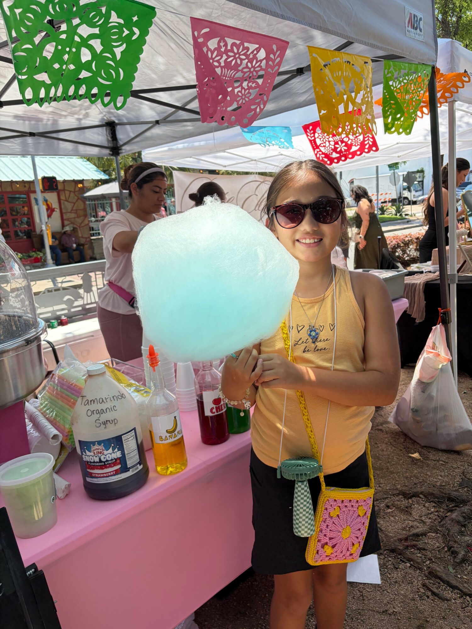 Girl with sunglasses holding cotton candy at outdoor event, pink table with drinks and decorations, sunny day.