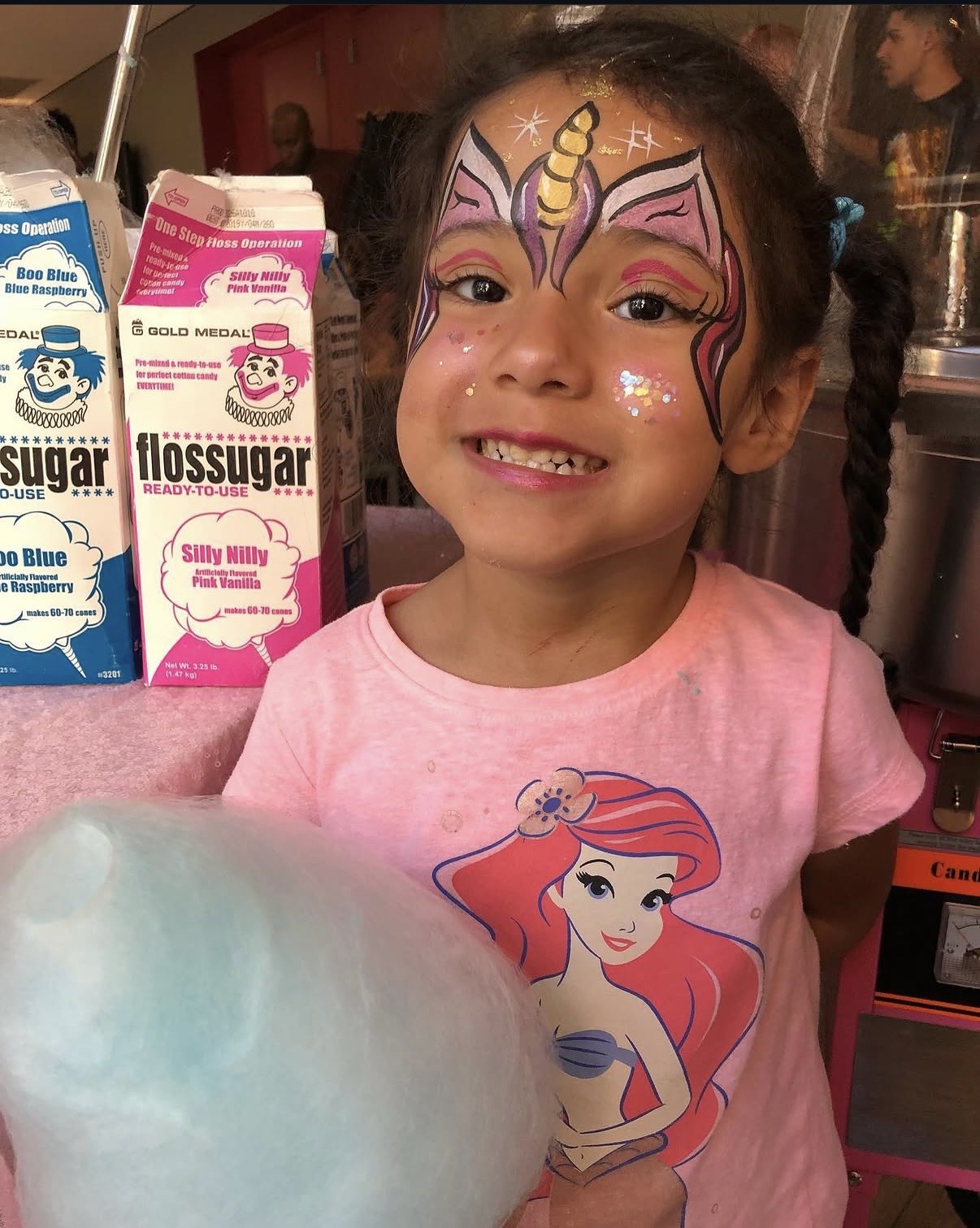 Young girl with face paint of a unicorn, smiling, wearing a pink shirt with a mermaid illustration, standing indoors.