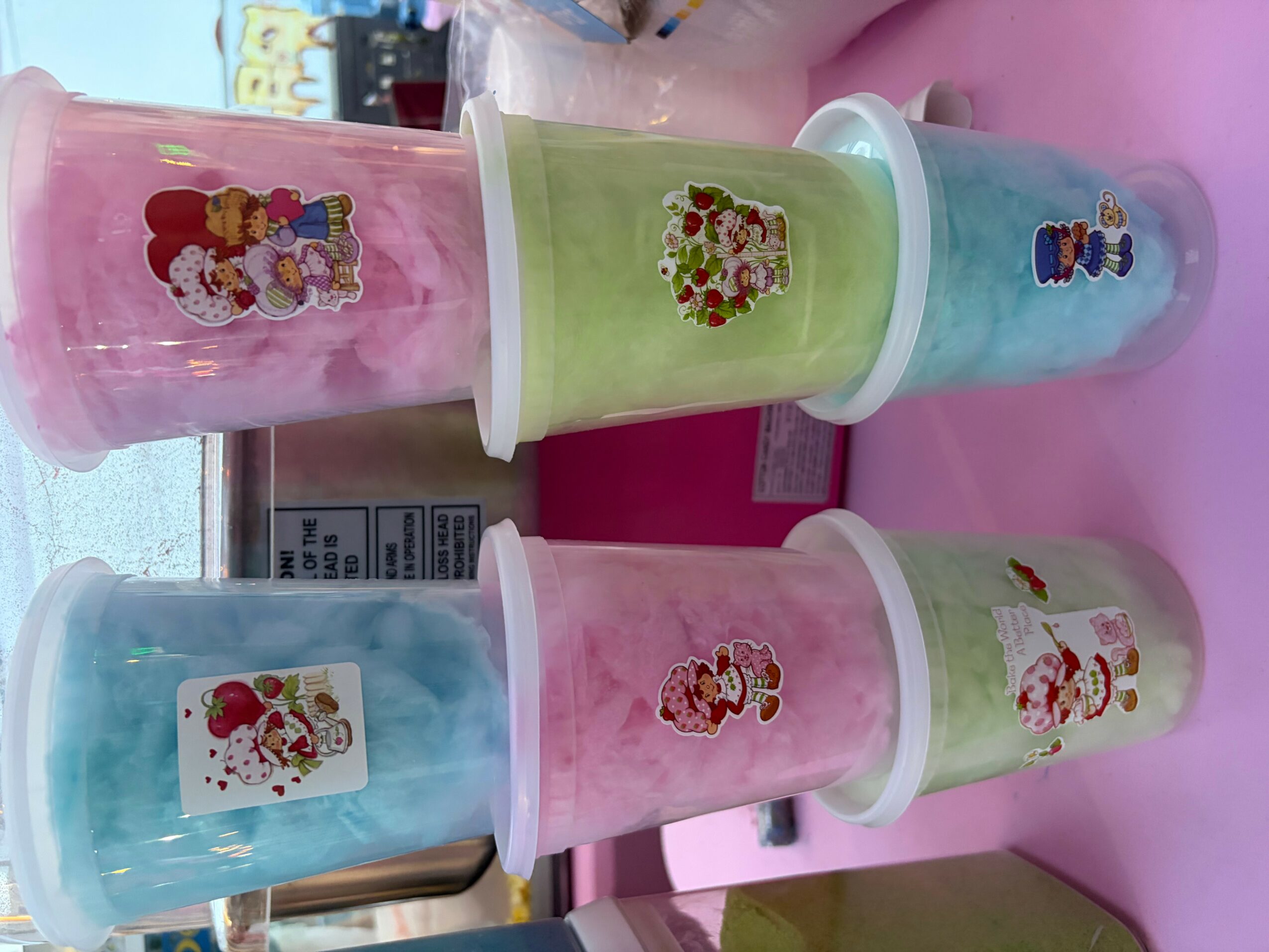 Six colorful cups with cartoon character stickers, arranged on a pink surface, with a window and objects in the background.
