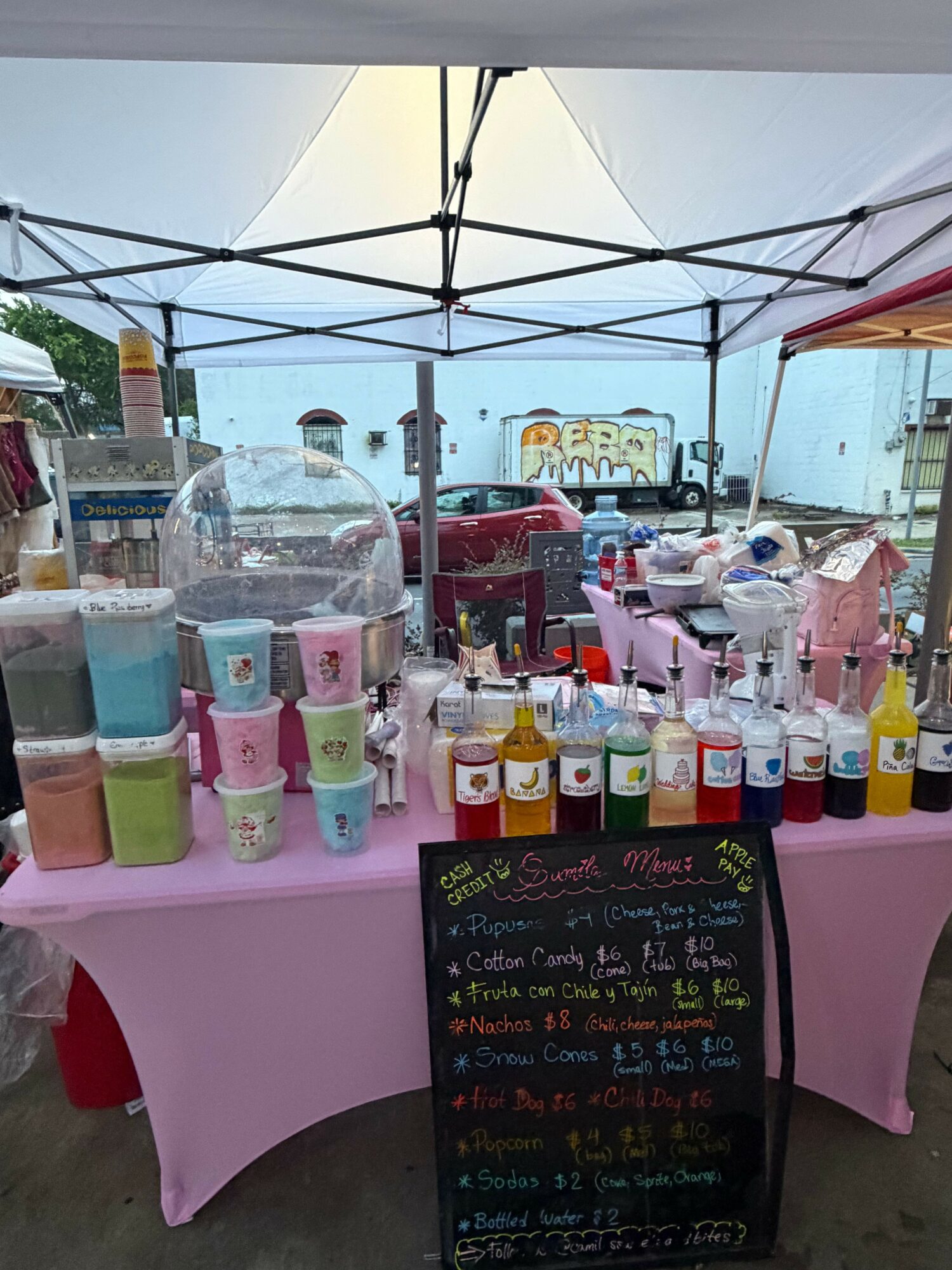 Display of colorful bottles and a chalkboard menu at a market stall with pink tablecloth.