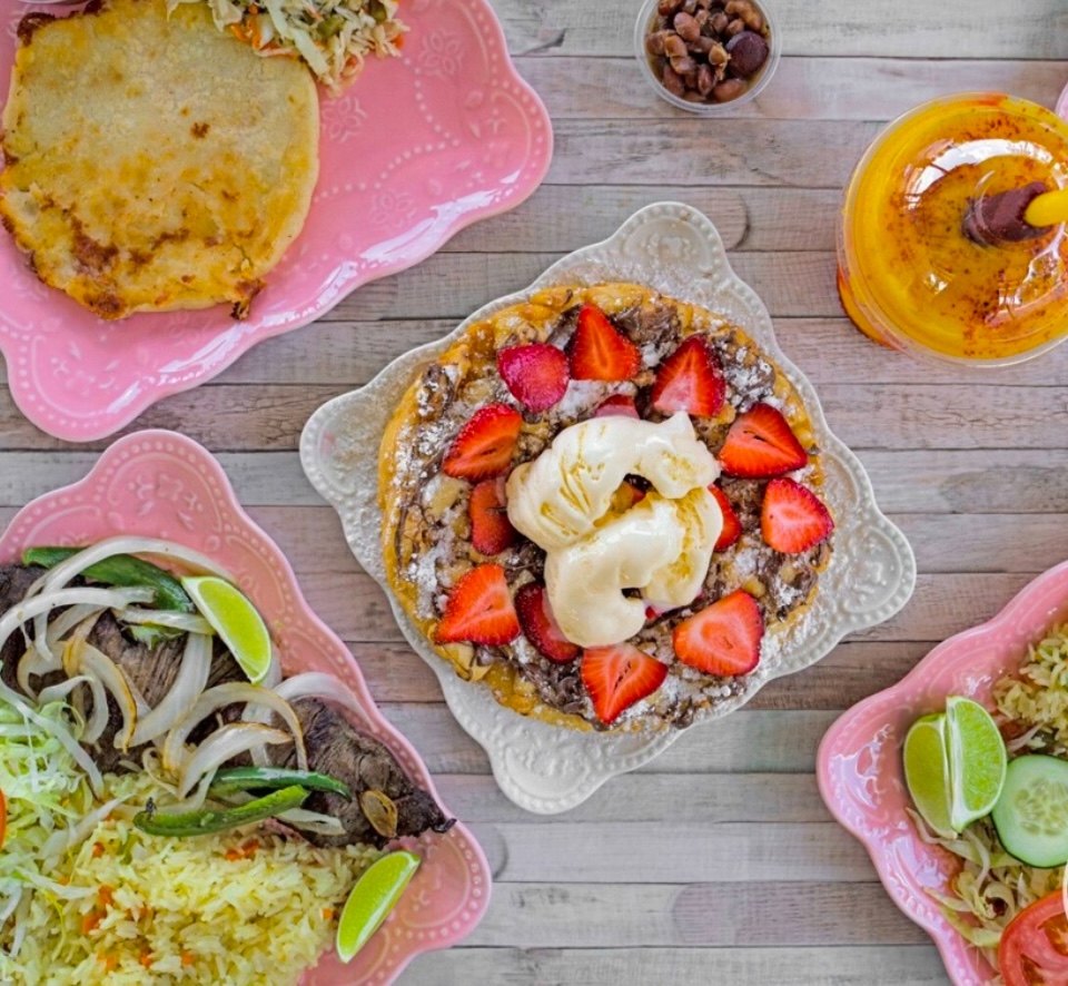Dessert topped with strawberries, banana slices, and ice cream on a white plate, surrounded by other dishes and drinks on a wooden table.