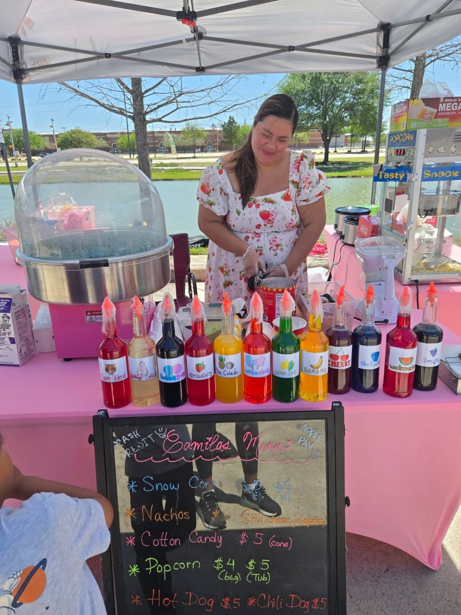 Woman standing behind a pink cotton candy stand with colorful bottles and a cotton candy machine outdoors.