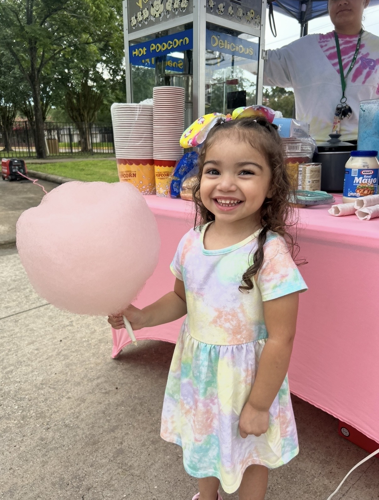 Smiling girl holding pink cotton candy at outdoor event, wearing pastel dress and colorful headband, with vendor stand in background.