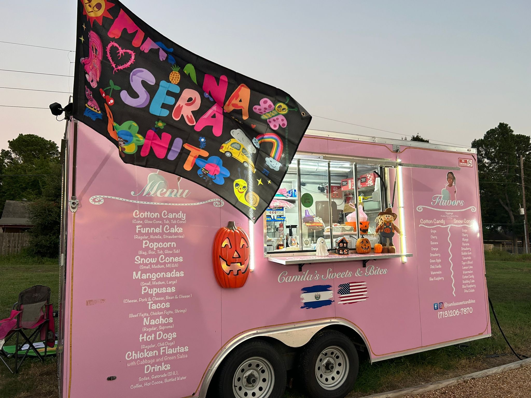Pink food truck with a colorful sign, menu, and a Halloween pumpkin decoration, parked outdoors with trees in the background.