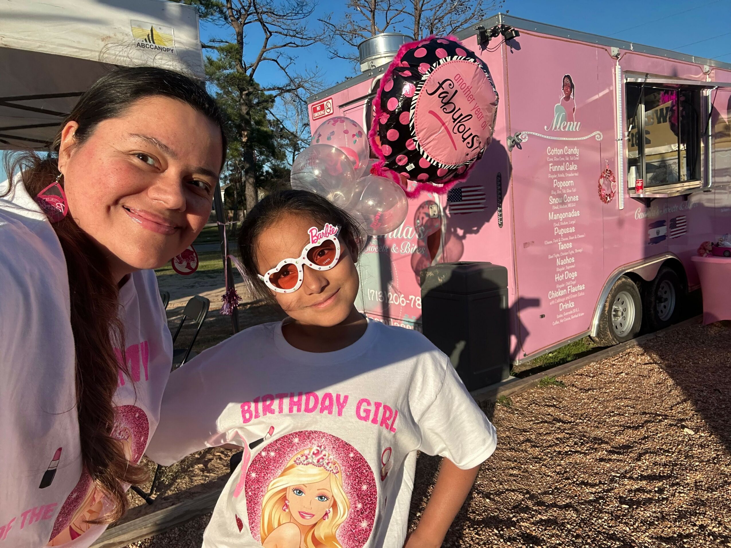 Two people, one child wearing sunglasses and a pink hat, and an adult, are outdoors near a pink food truck.
