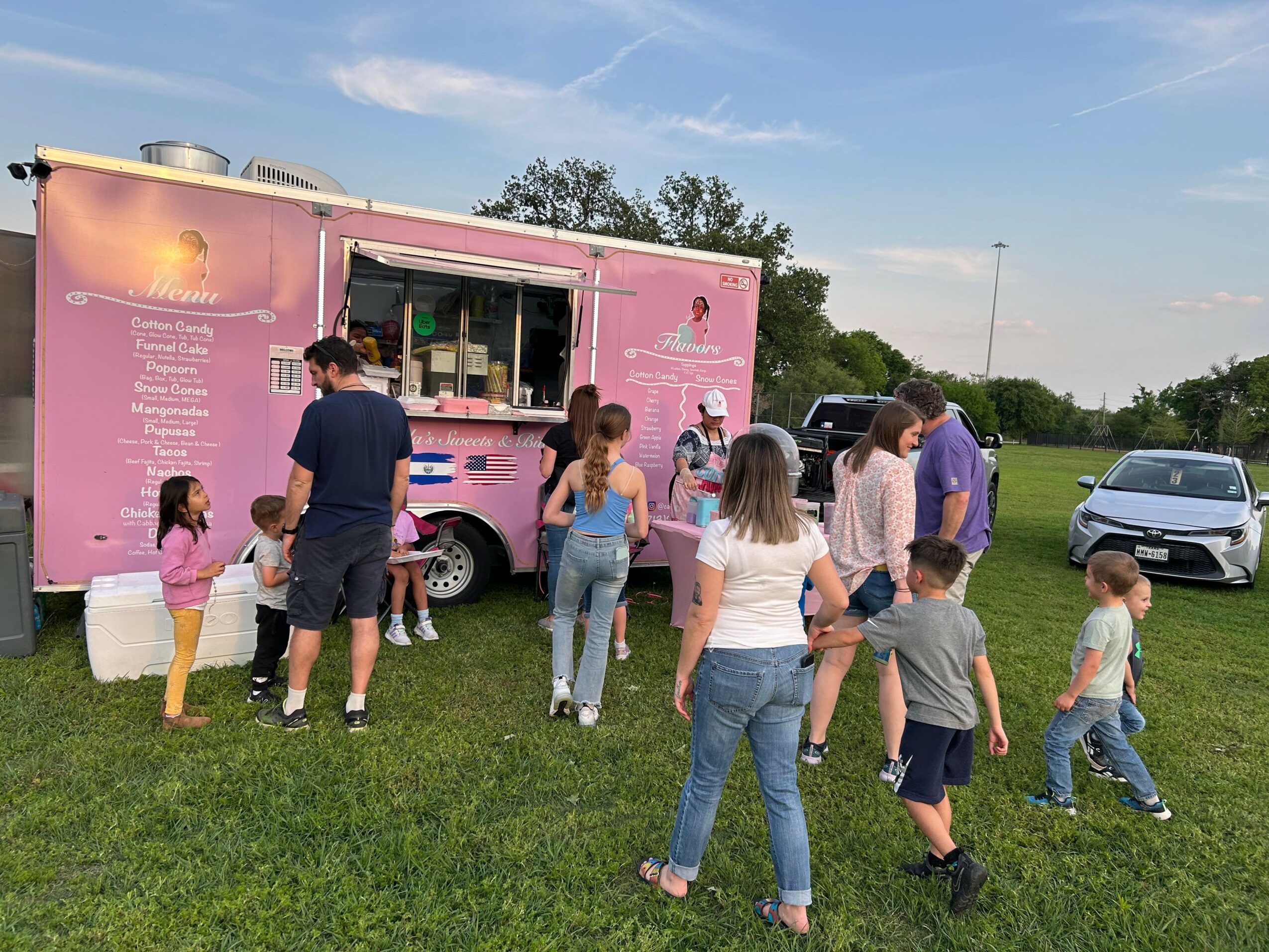 People line up at a pink food truck outdoors on a grassy field, with a car and trees in the background.